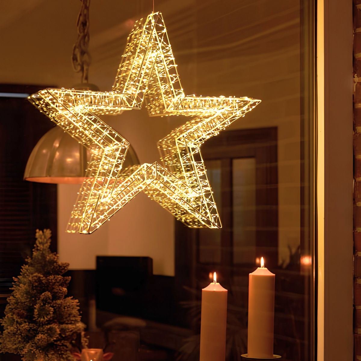 Een oplichtende draadster hanglamp, twee brandende kaarsen en een mini kerstboom in een kamer.