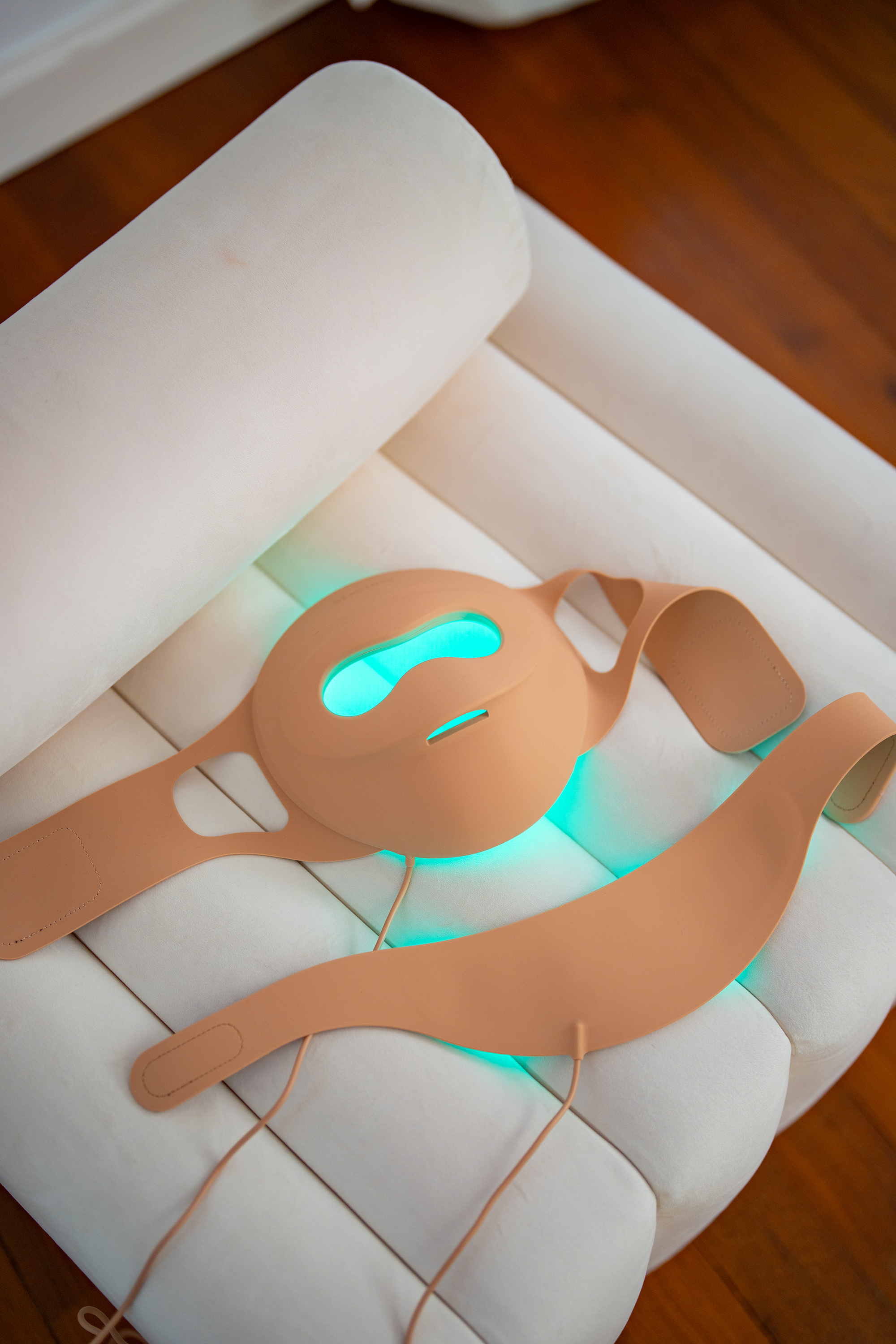 Una maschera facciale color carne con illuminazione LED verde brillante giace su un divano bianco.