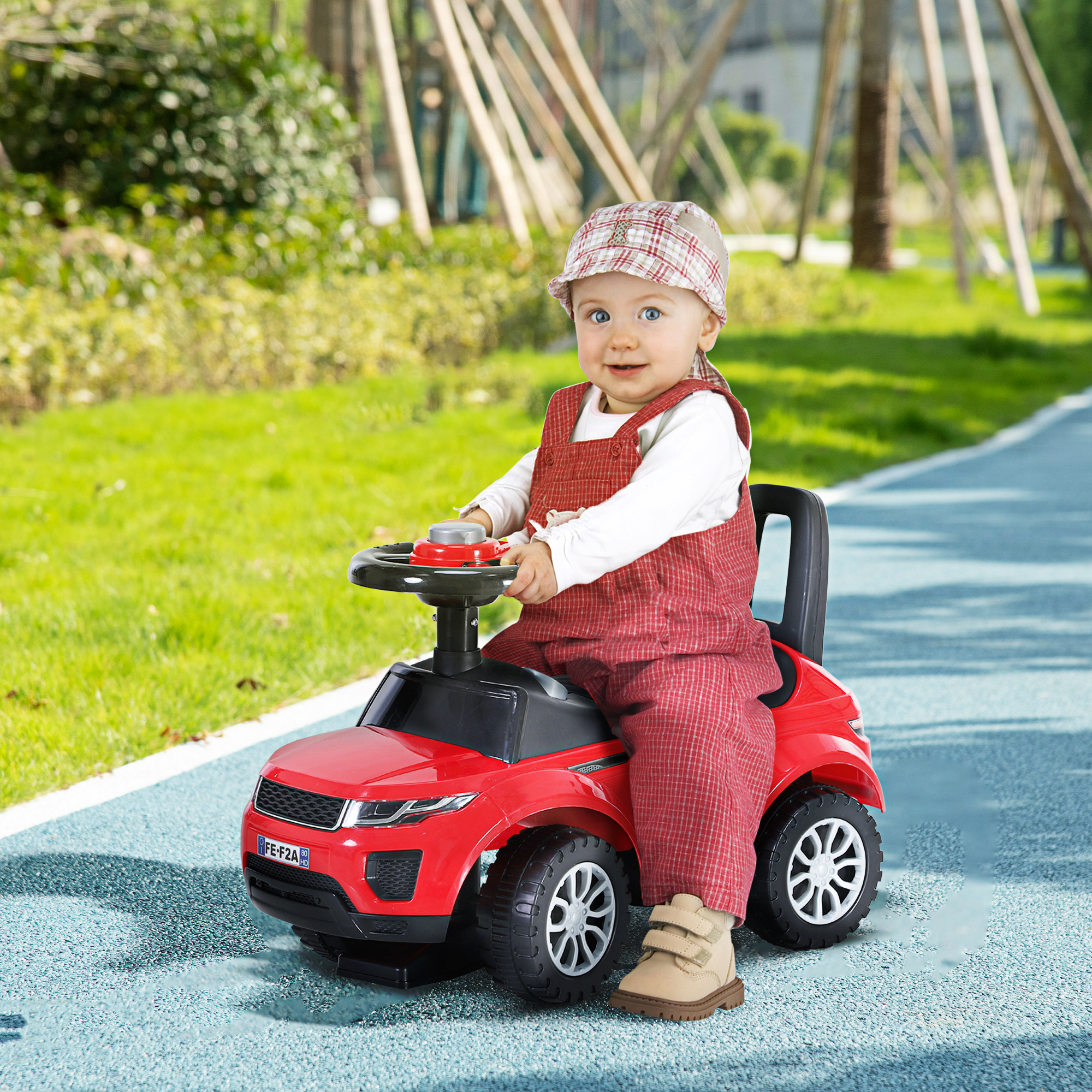Bambino piccolo con salopette rossa guida un'auto giocattolo rossa su un sentiero blu in un parco.