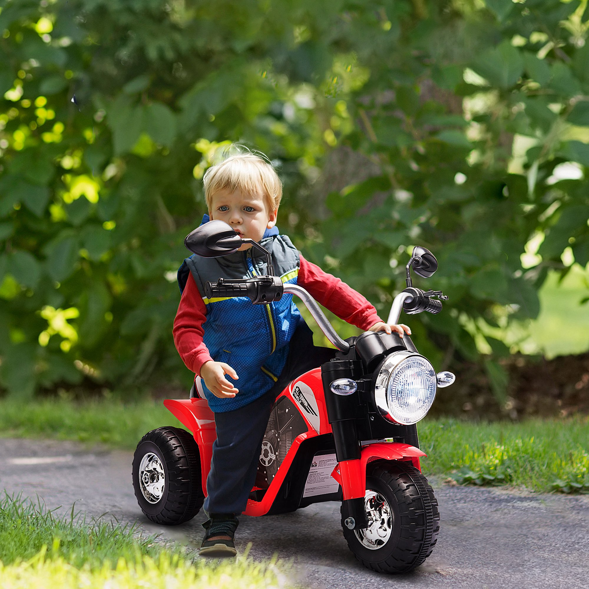 Un bambino guida una motocicletta giocattolo rossa a tre ruote all'aperto su un sentiero asfaltato, circondato da verde.