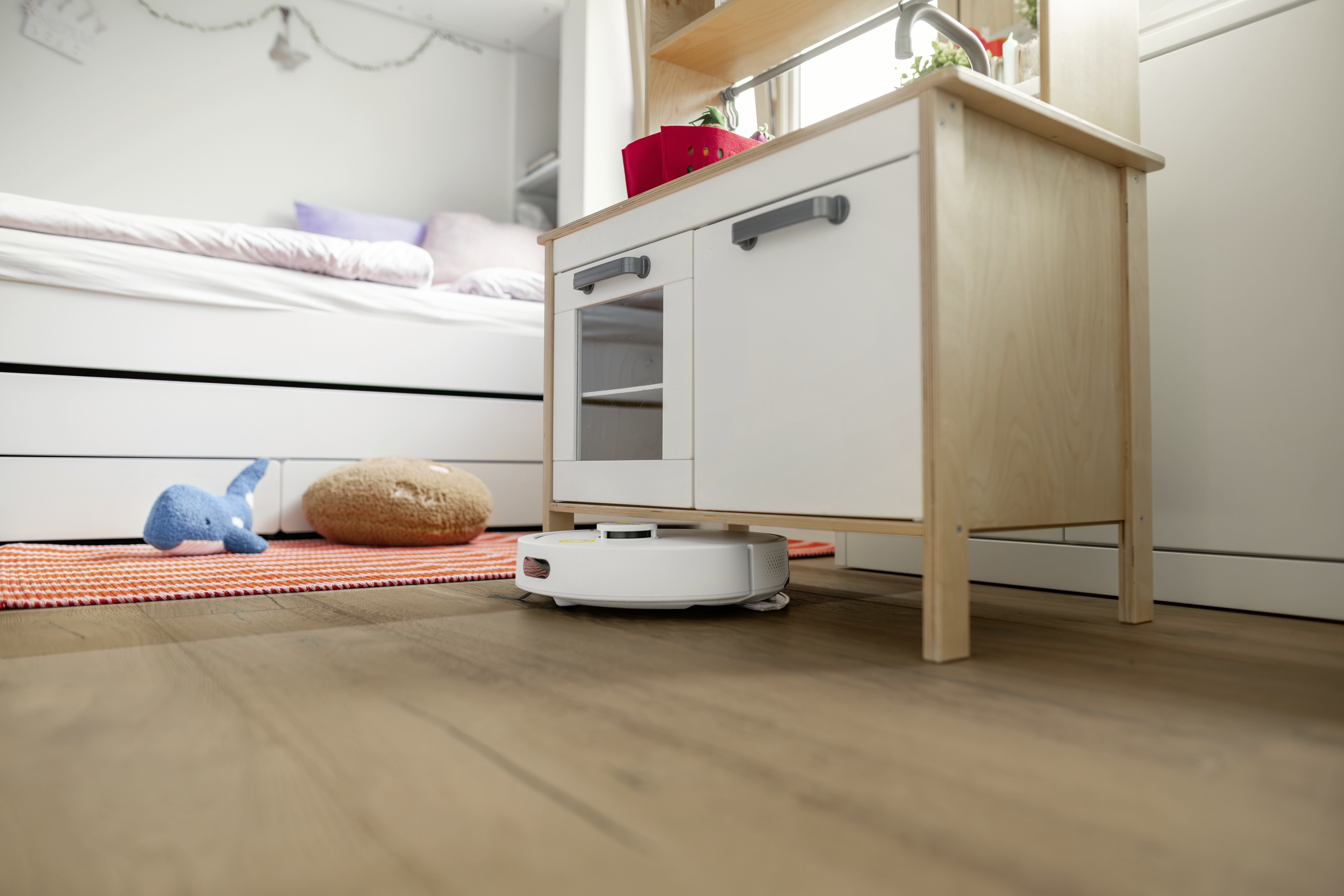 A white robot vacuum cleaner cleans the wooden floor in a children's room under a play kitchen.