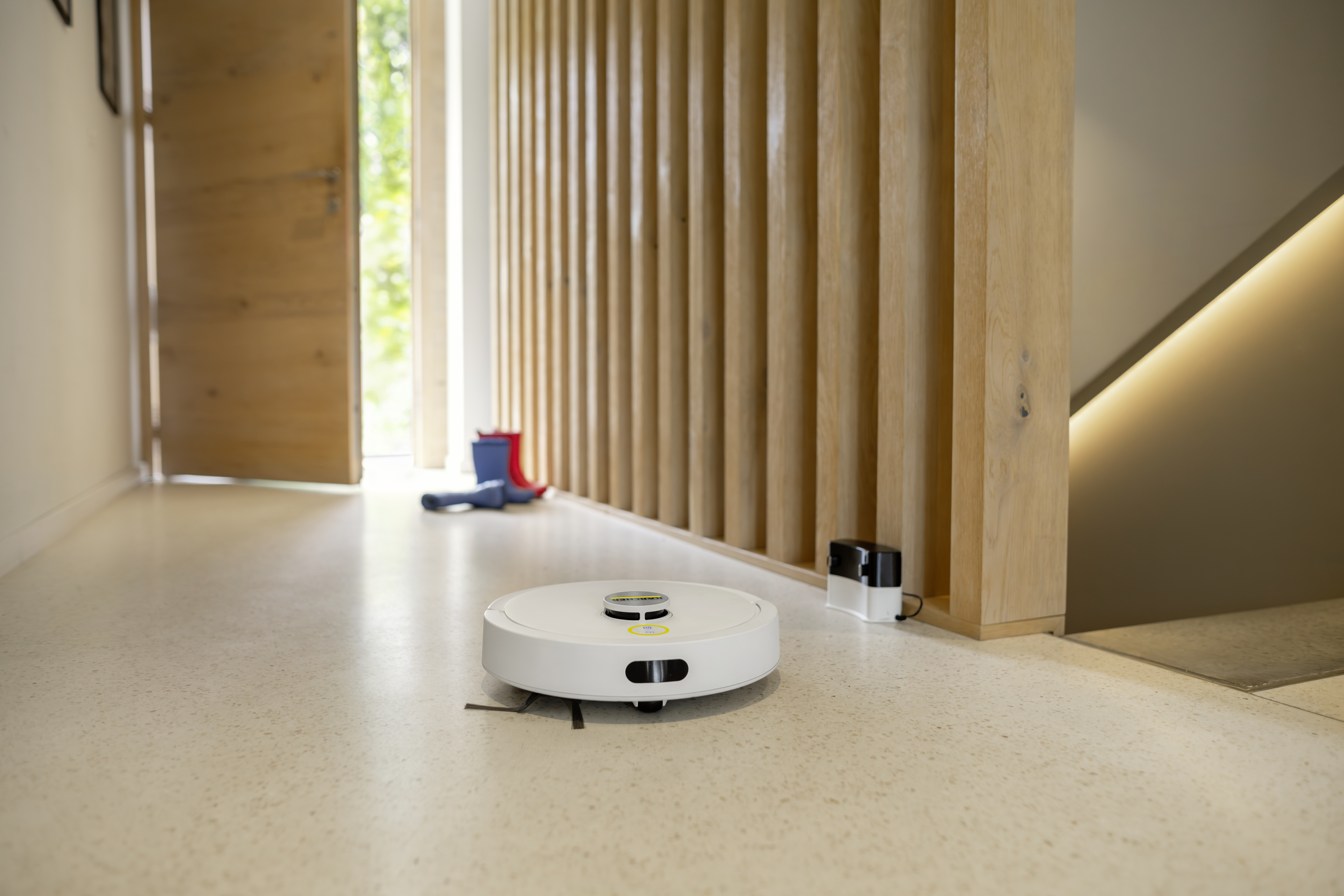 A white robot vacuum cleaner sits on the bright floor of a modern hallway with wooden wall paneling.