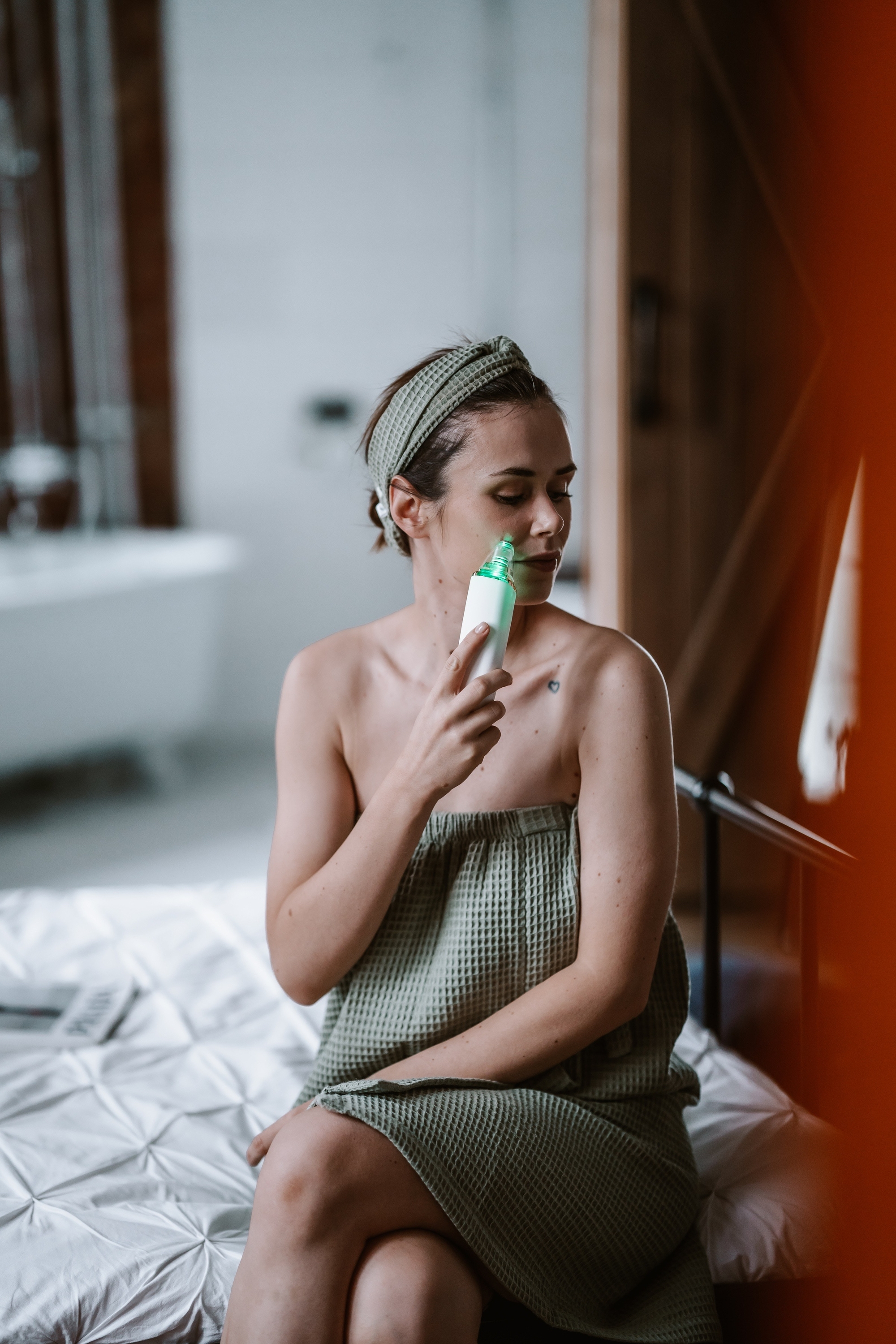 Una donna con asciugamano verde e fascia usa un dispositivo per il viso sul letto.