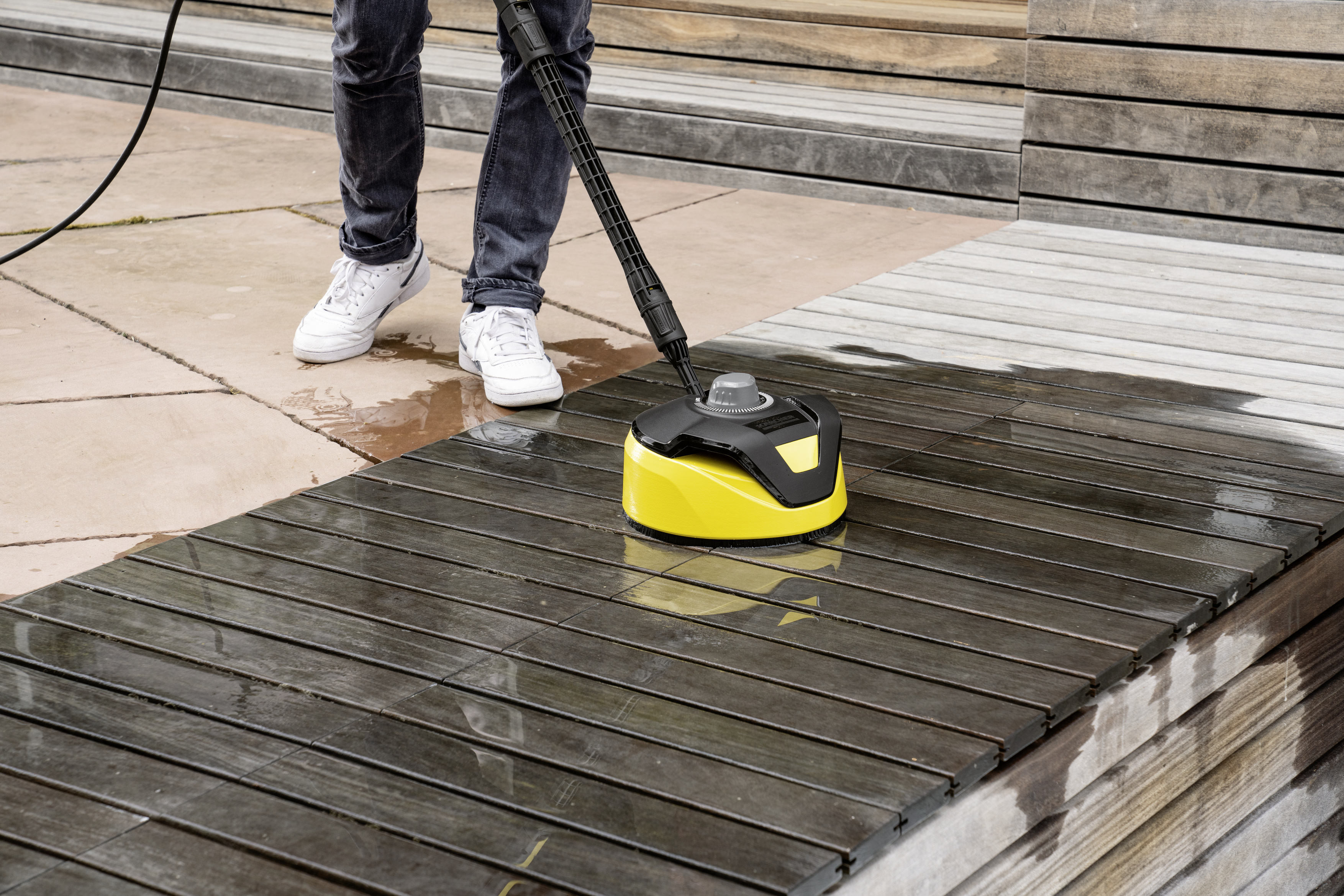 Eine Person reinigt mit einem gelb-schwarzen Flächenreiniger ein Holzdeck.