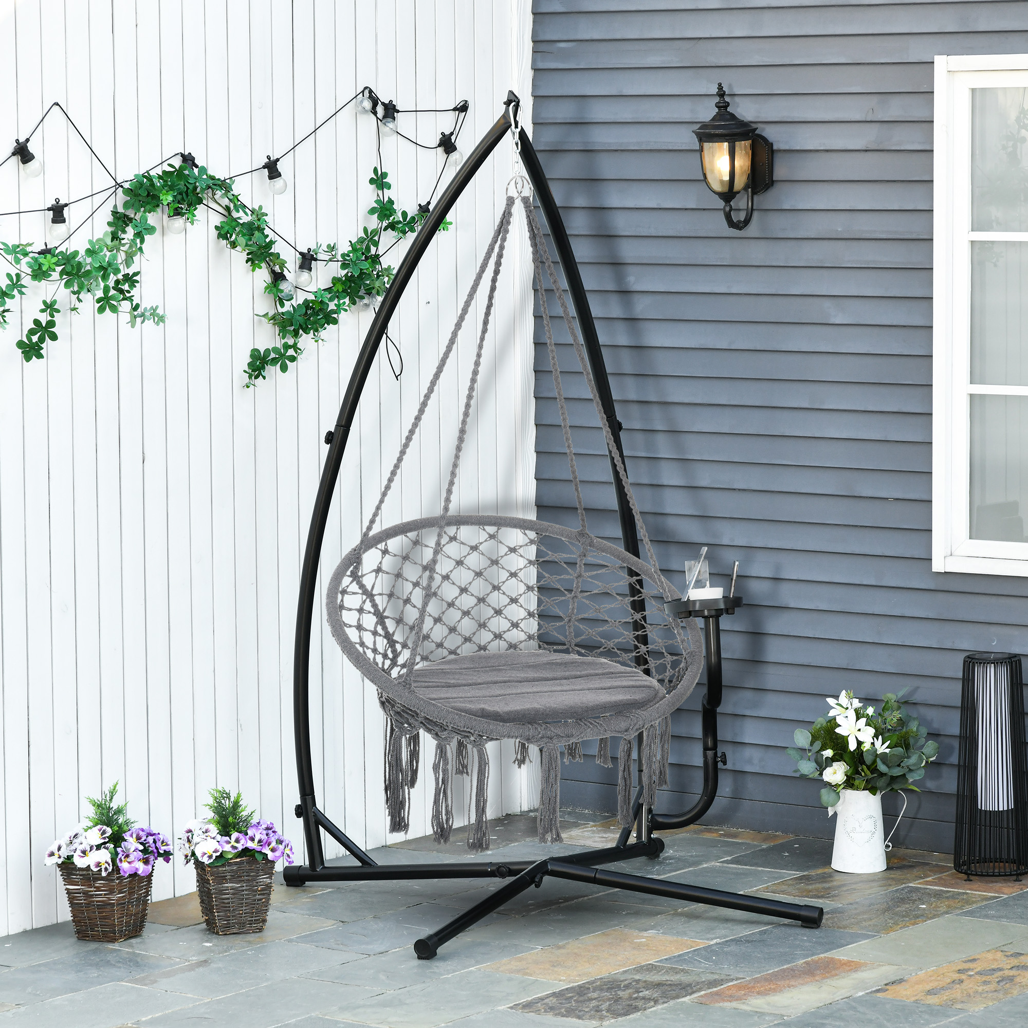 Una silla colgante de macramé gris con soporte metálico negro en un patio, flanqueada por macetas y luces.