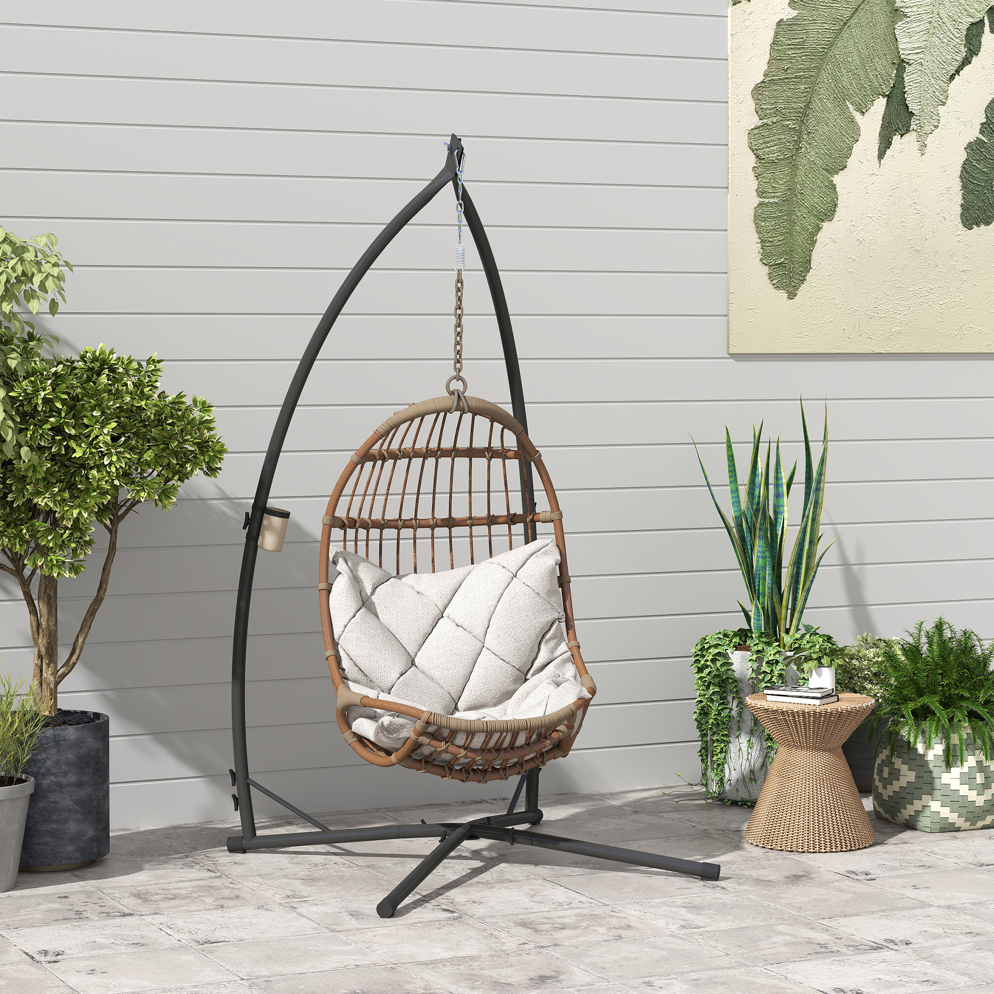 Una silla colgante de mimbre con cojín blanco en soporte negro, rodeada de plantas en macetas en un patio.