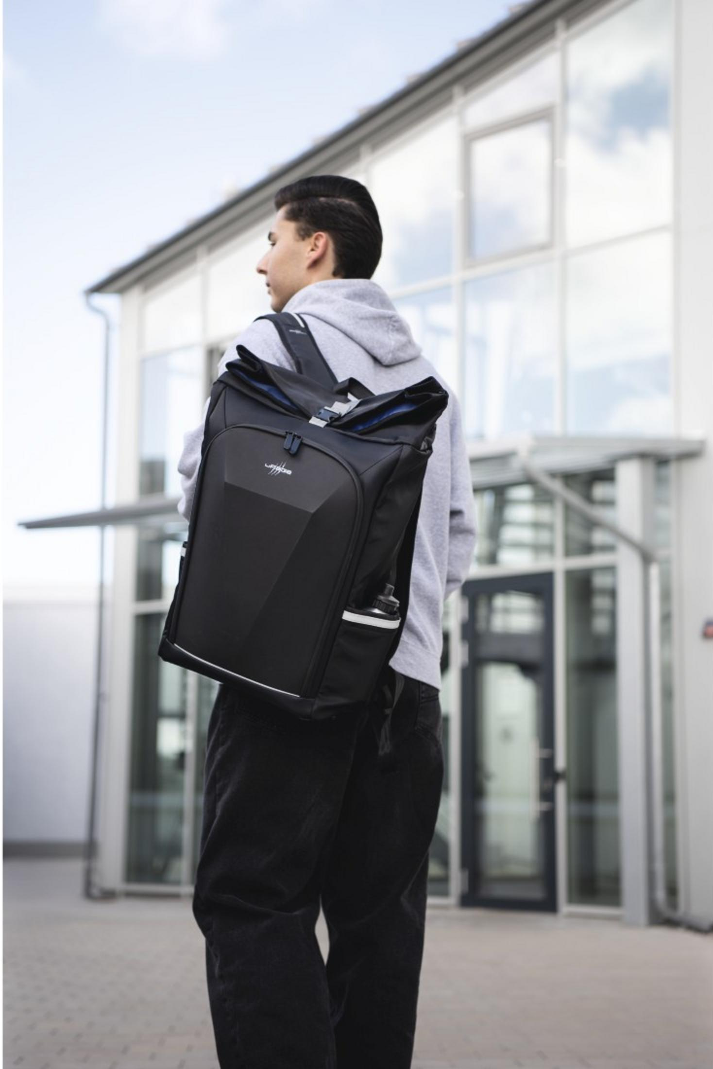 Ein junger Mann mit grauem Hoodie und schwarzem Rucksack steht vor einem modernen Gebäude.