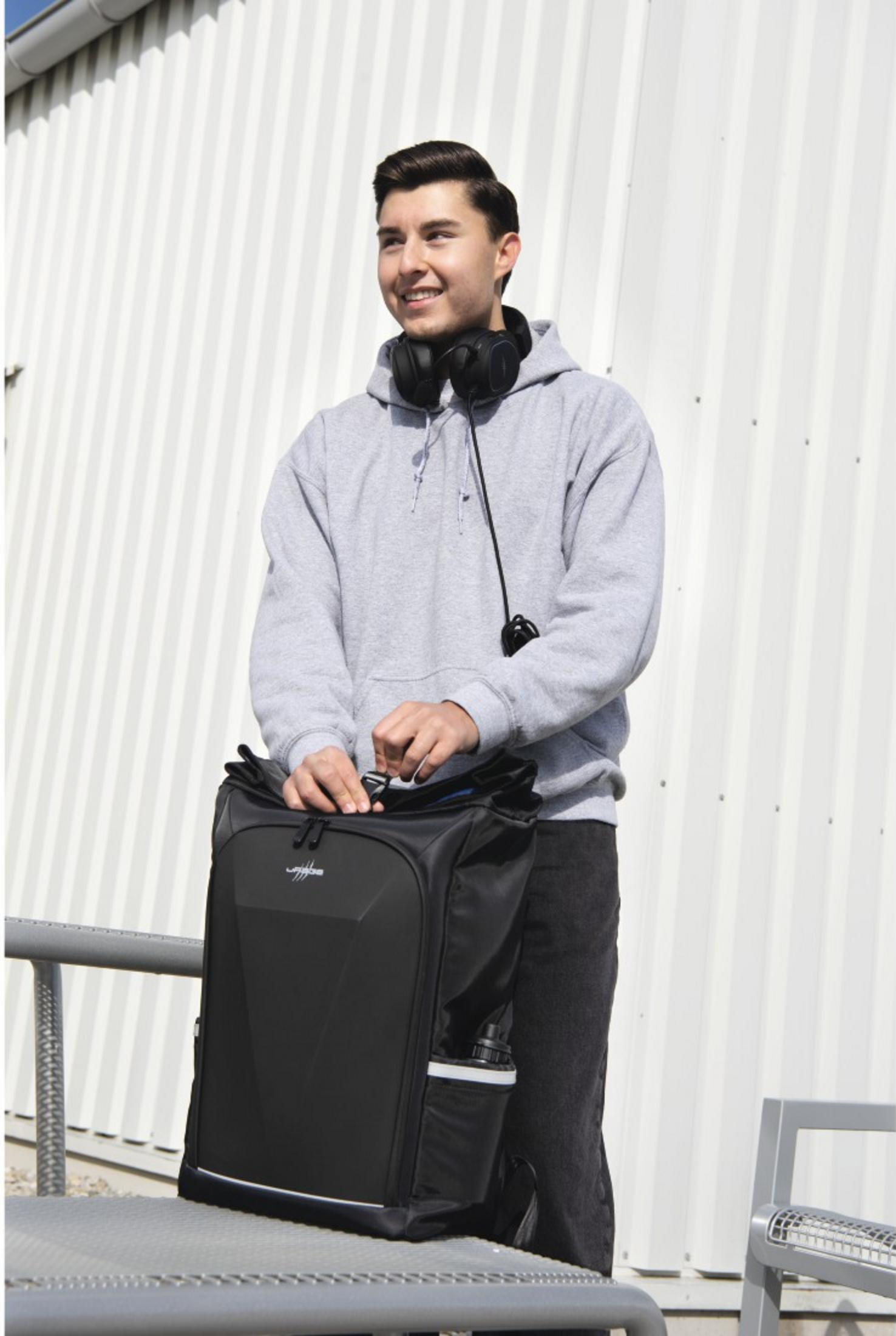 Ein junger Mann mit Graublatt-Hoodie und Kopfhörern öffnet einen großen schwarzen Rucksack im Freien vor einer weißen Wand.