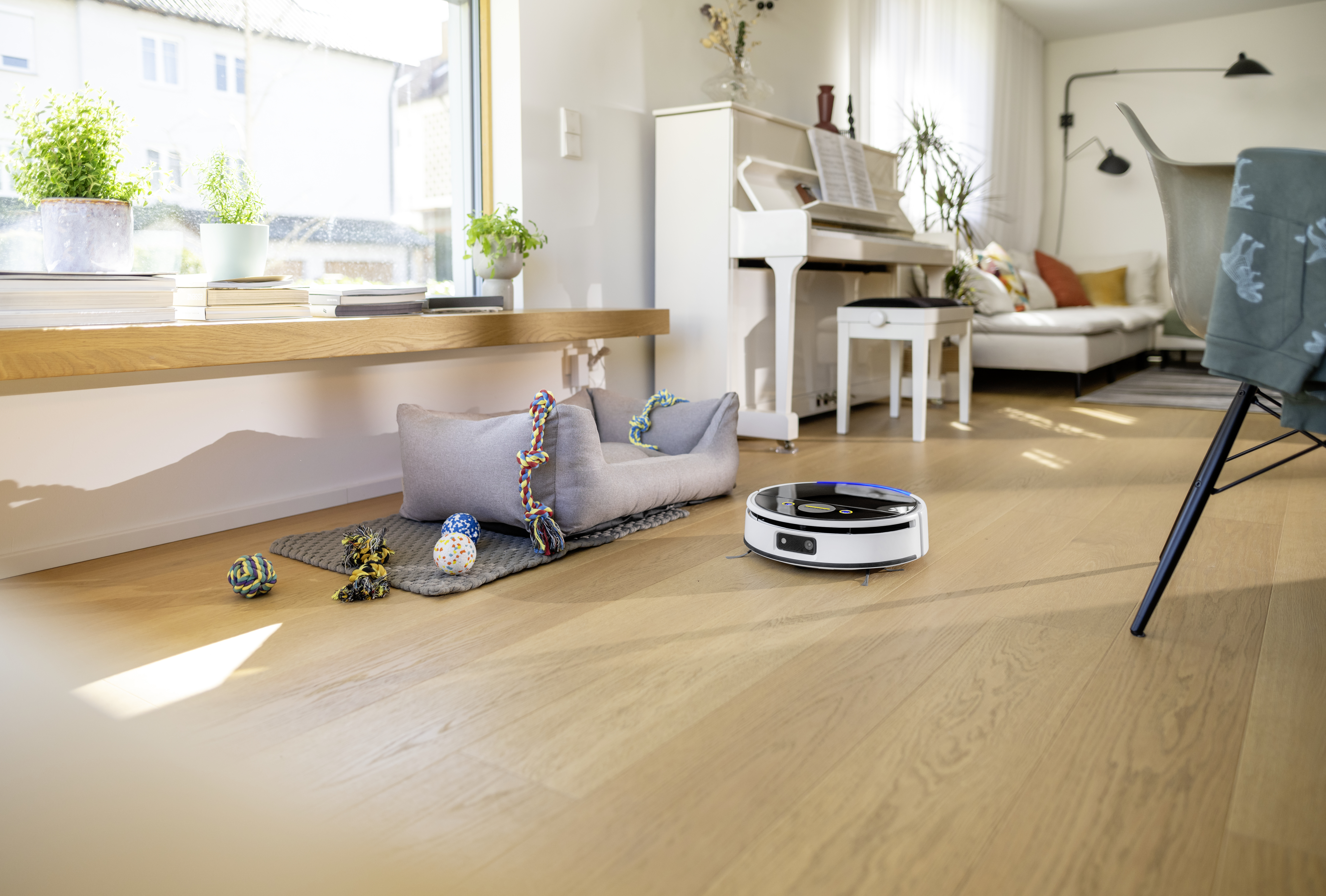 Un robot aspirapolvere su un pavimento in legno in un soggiorno, vicino a una cuccia grigia e giocattoli.