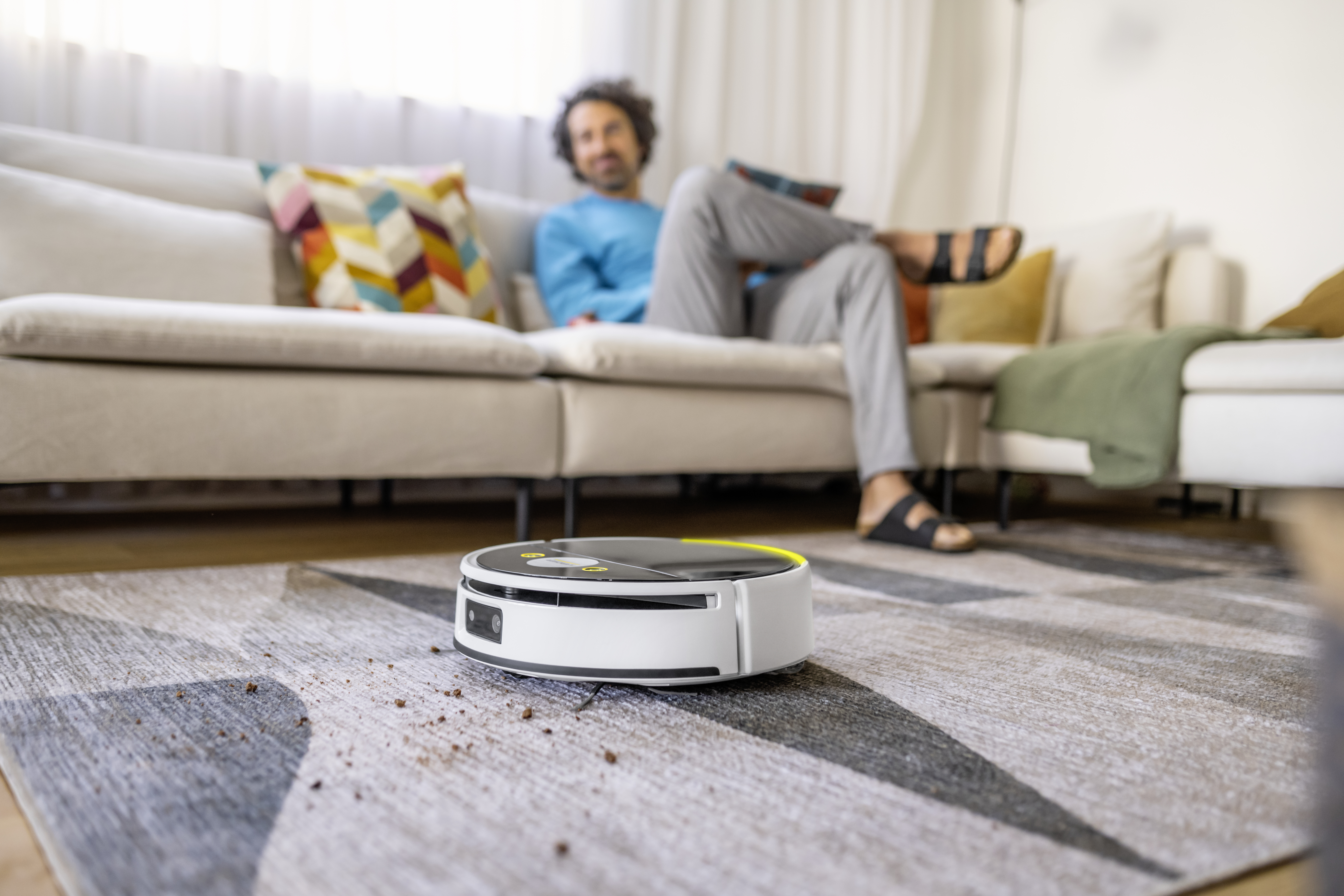 Un robot aspirapolvere pulisce un tappeto decorato in un soggiorno mentre un uomo si rilassa sul divano.