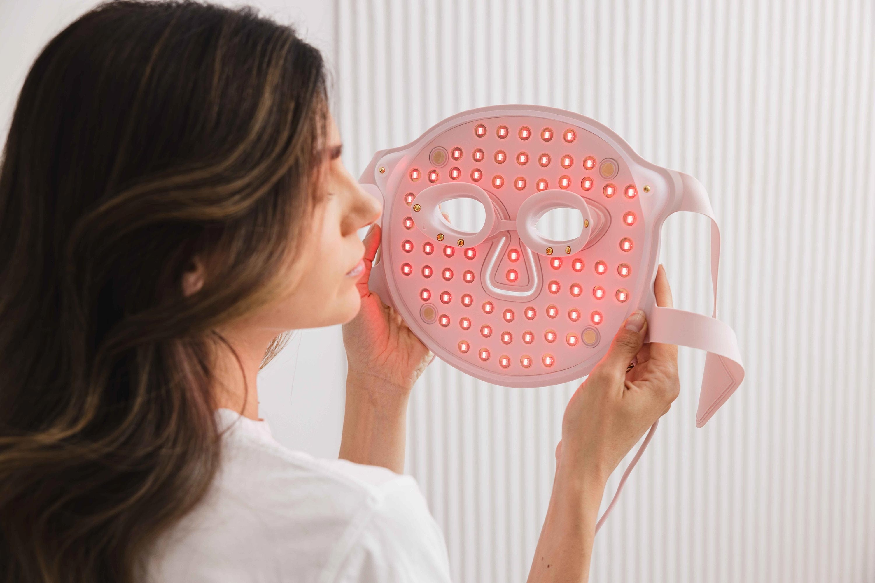 Una donna tiene in mano una maschera facciale a LED rosa con luce rossa brillante davanti al viso.