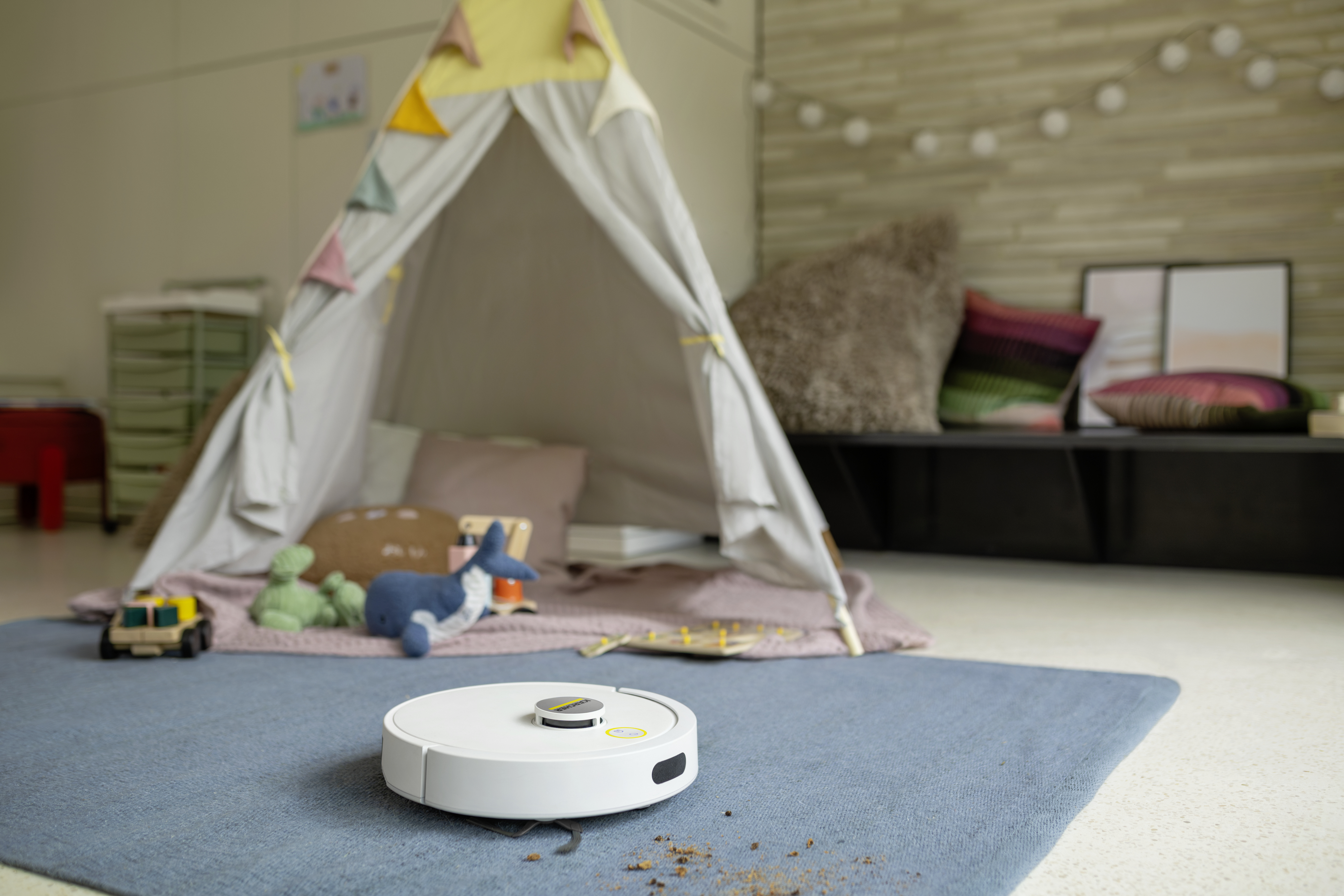 Ein weißer Saugroboter reinigt einen blauen Teppich in einem gemütlichen Kinderzimmer vor einem Spielzelt.