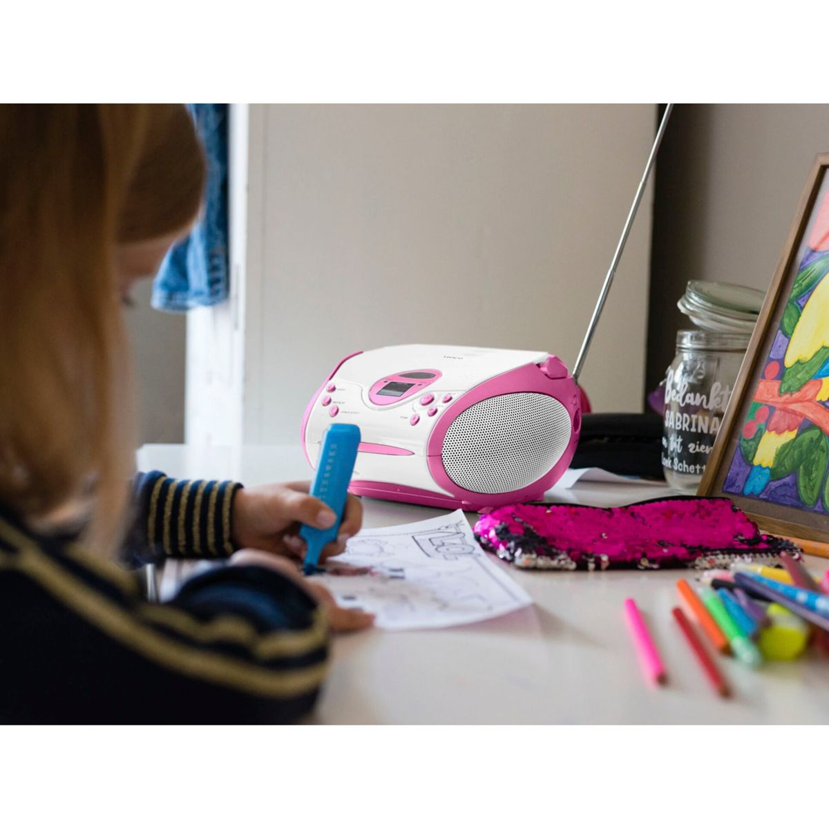 Una ragazza disegna a un tavolo con uno stereo portatile rosa e bianco vicino.