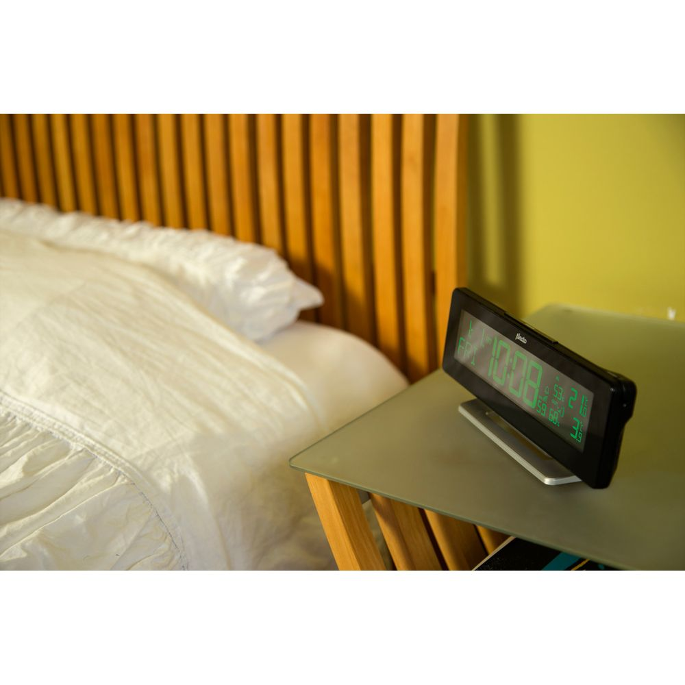 Un réveil numérique à affichage vert est posé sur une table de chevet près d'un lit en bois.