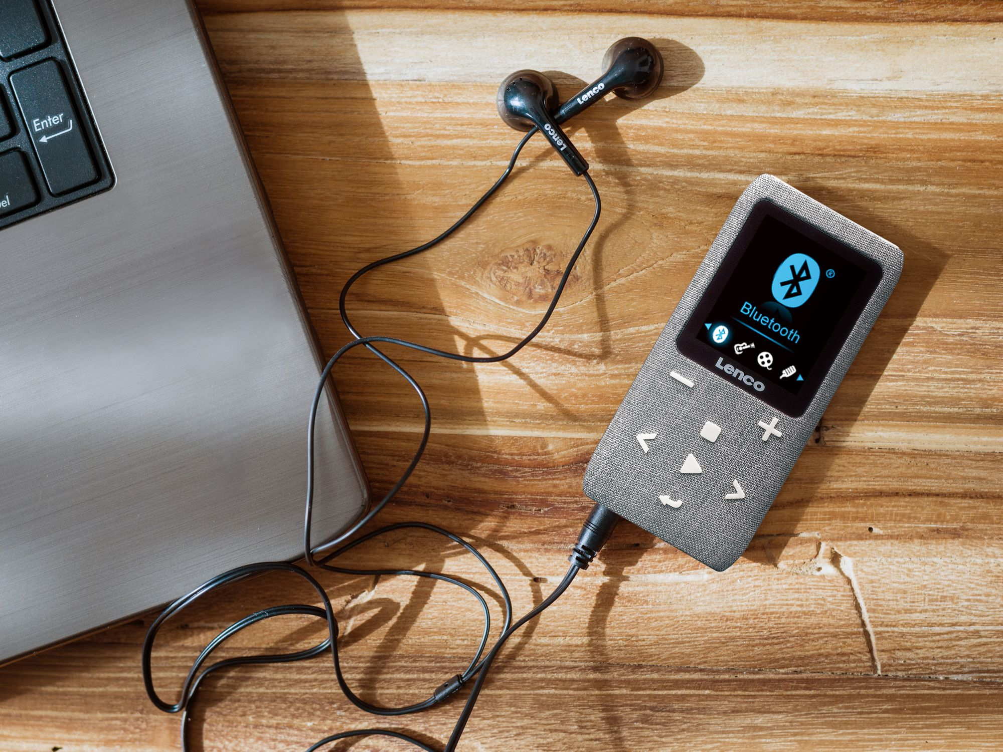 Un lettore MP3 Lenco grigio con auricolari e computer portatile su un tavolo di legno.