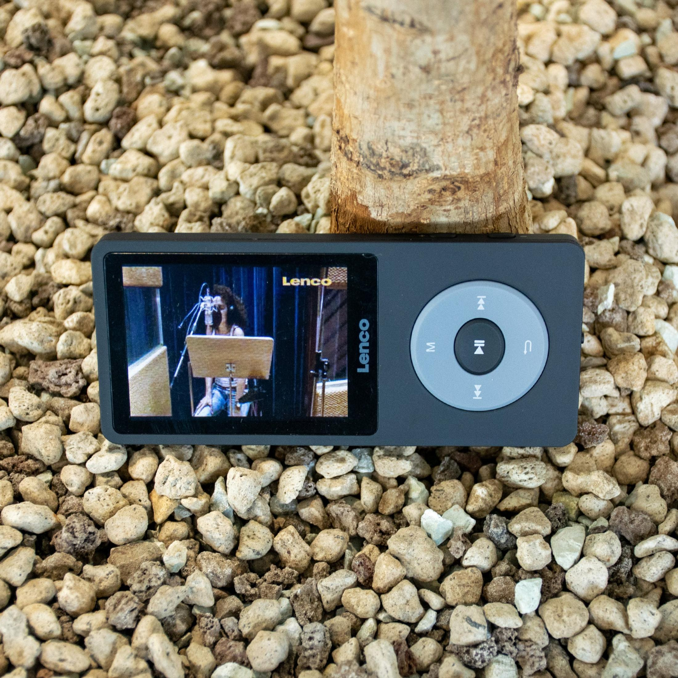 Ein schwarzer Lenco-Media-Player auf kleinen, hellen Steinen in der Nähe eines Baumstamms, der ein Video wiedergibt.