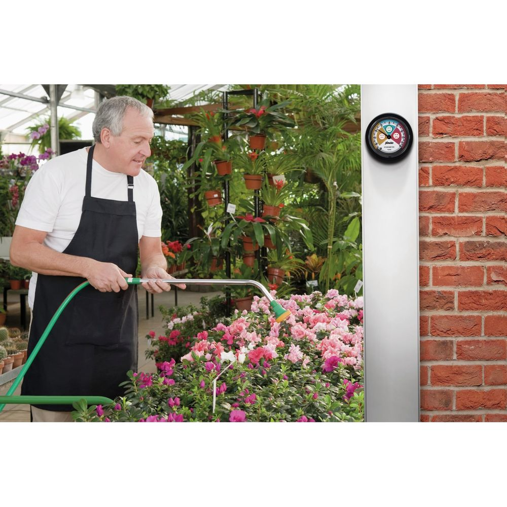 Ein Gärtner in schwarzer Schürze bewässert rosa Blumen in einem Gewächshaus, daneben eine Wand mit Thermometer.