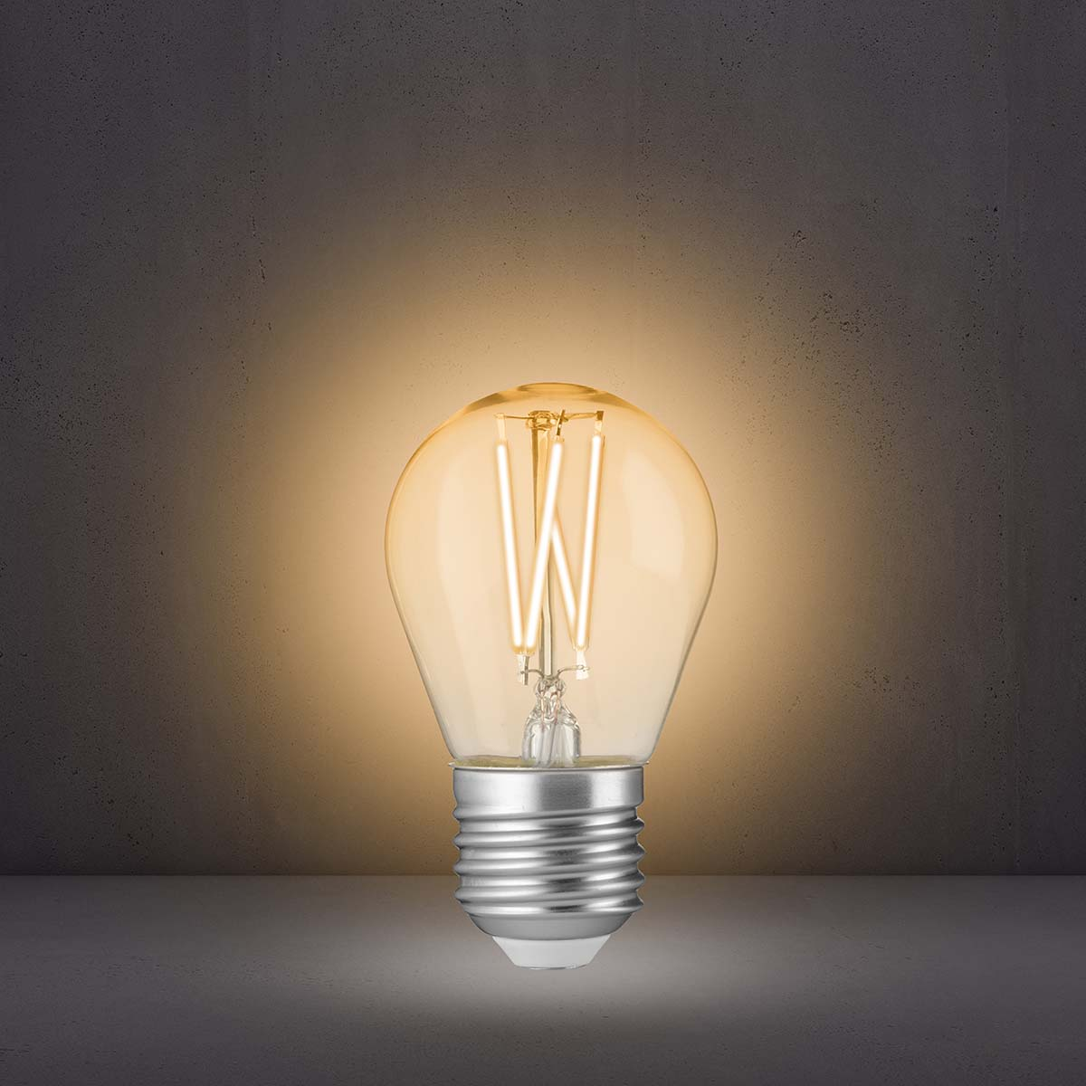 Eine leuchtende LED-Lampe mit sichtbaren Filamenten steht auf einer dunklen Fläche vor grauer Wand.