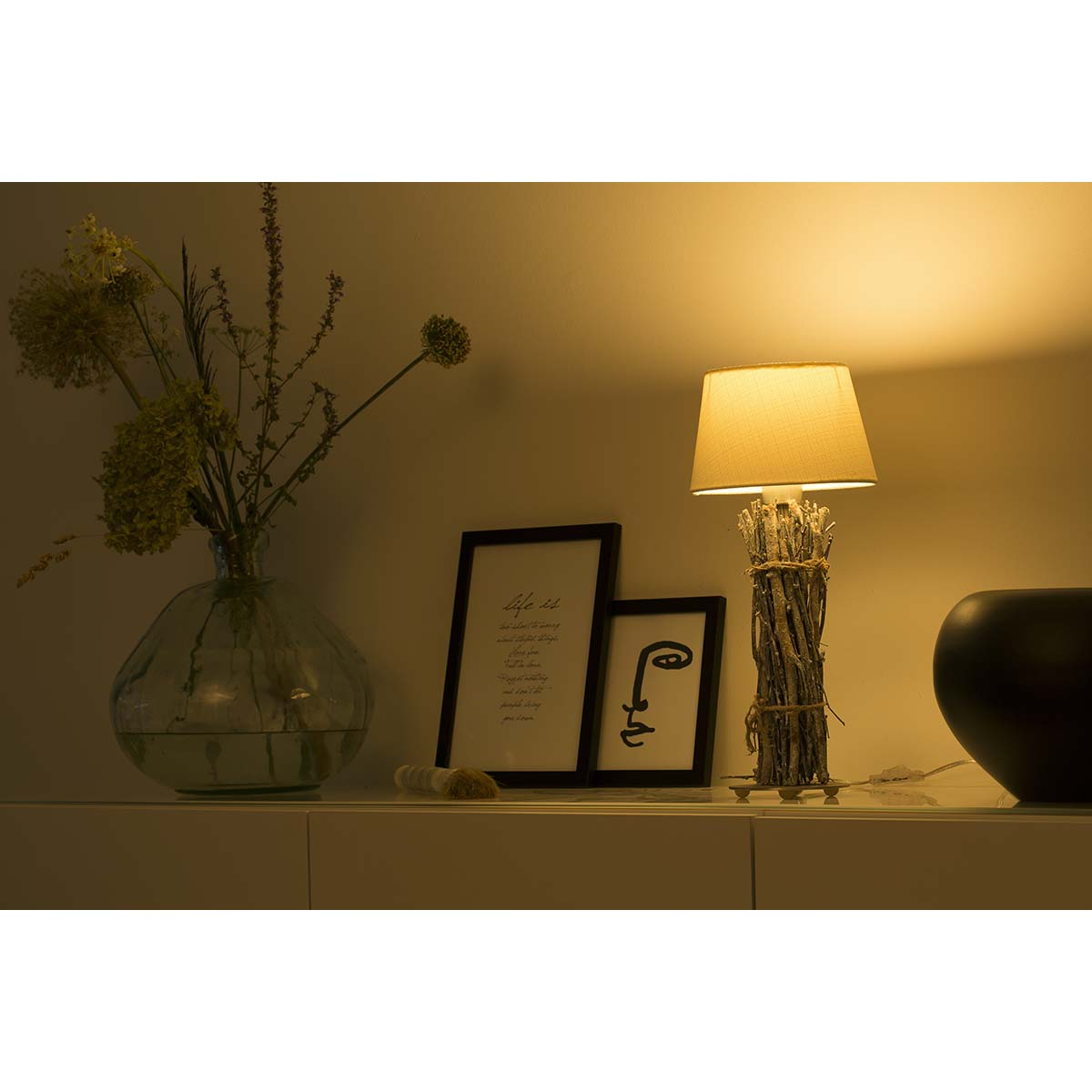 Eine beleuchtete rustikale Zweiglampe auf einem weißen Tisch neben gerahmter Kunst und einer Vase mit Trockenblumen.