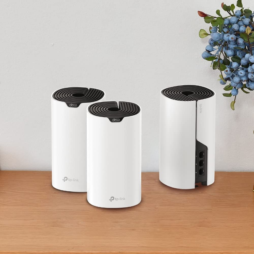 Tre router mesh TP-Link Deco bianchi cilindrici su un tavolo di legno contro una parete bianca con bacche blu artificiali.