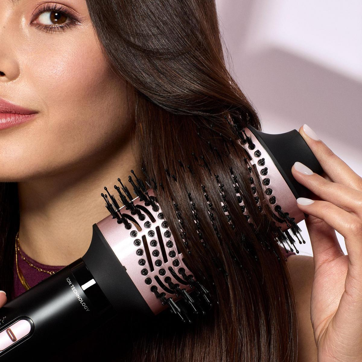 Una mujer se peina el pelo largo y castaño con un cepillo secador negro y rosa.