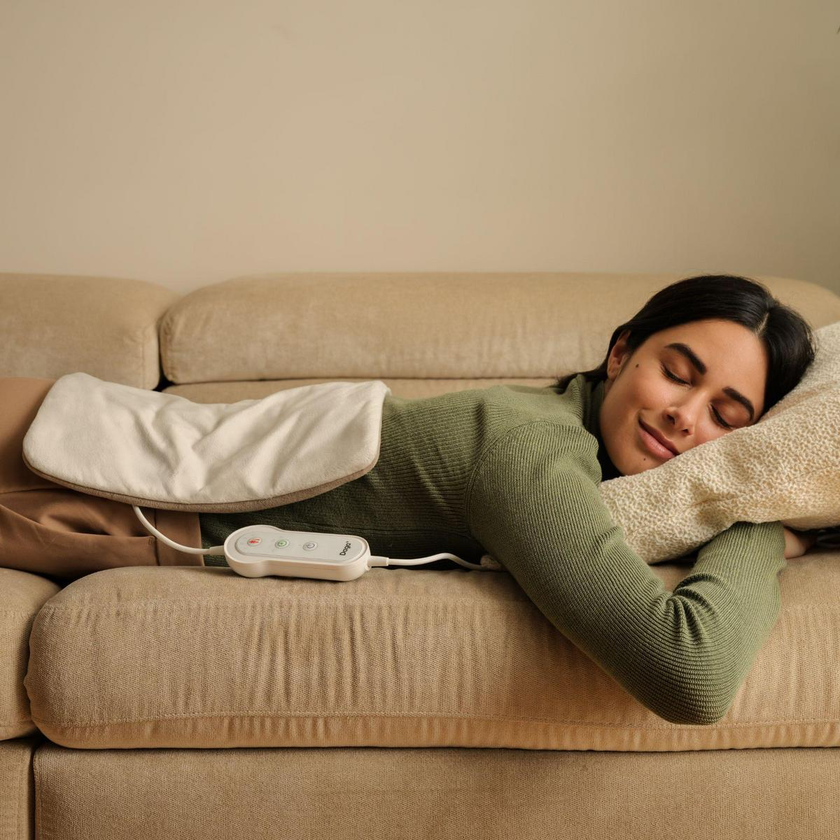 Una mujer duerme en un sofá beis con una almohadilla térmica en la espalda.