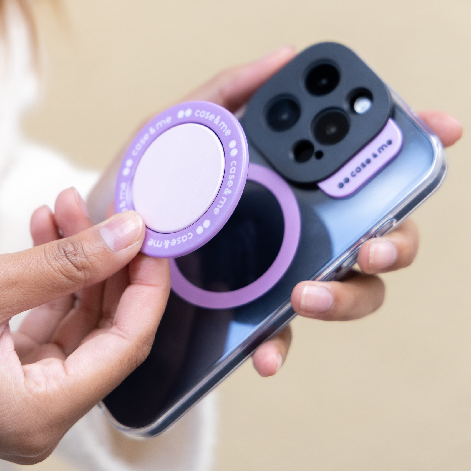 Manos sosteniendo un smartphone con funda transparente, protector de cámara morado y un anillo magnético morado a juego.