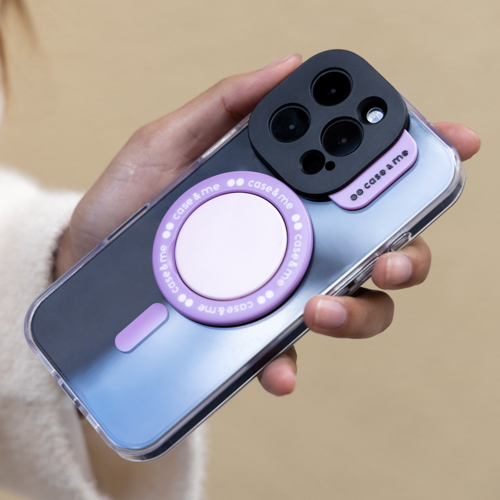 Mano sostiene smartphone transparente con módulo de cámara negro y anillo MagSafe morado.