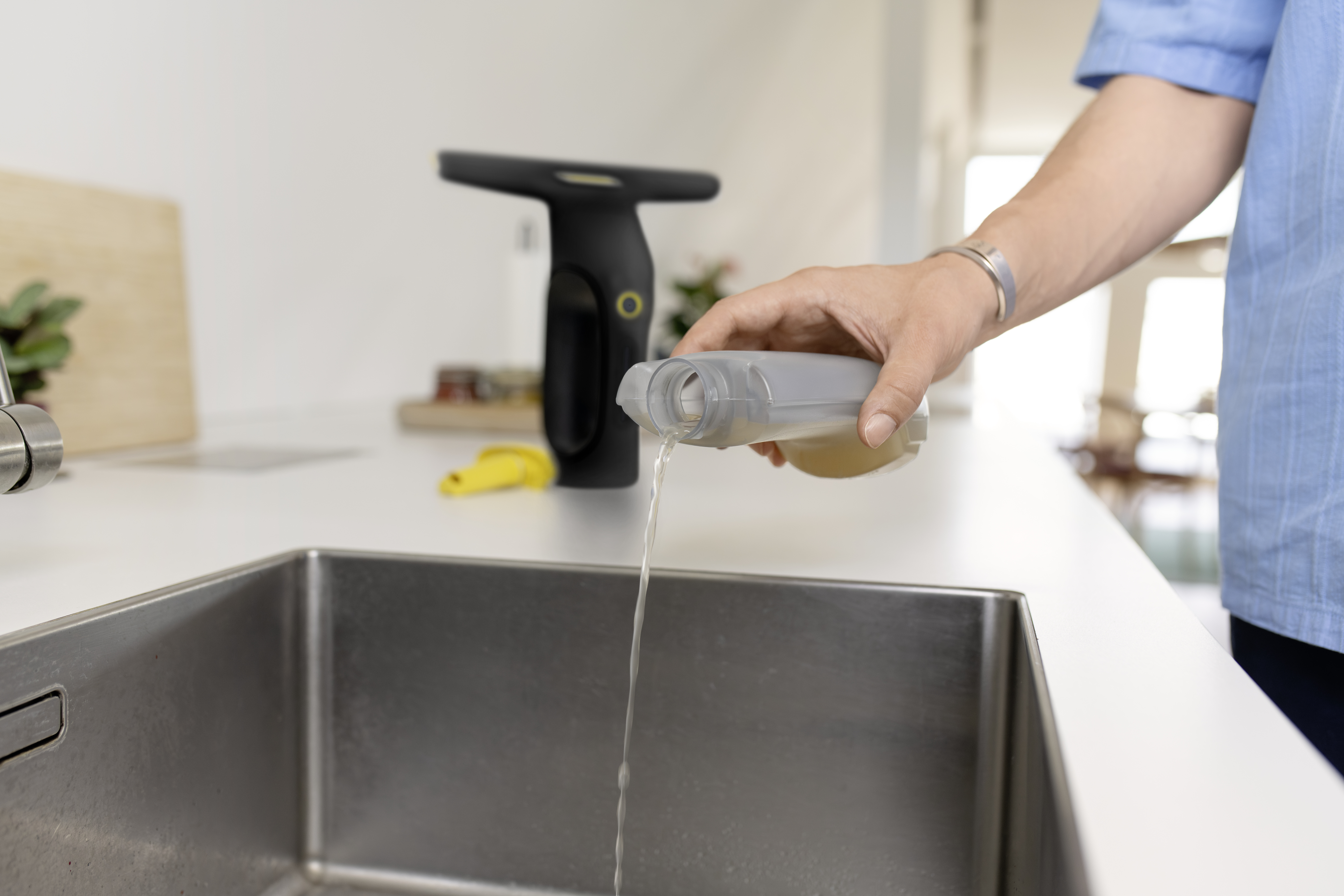 Eine Person gießt schmutziges Wasser aus einem grauen Behälter in ein Edelstahlwaschbecken.