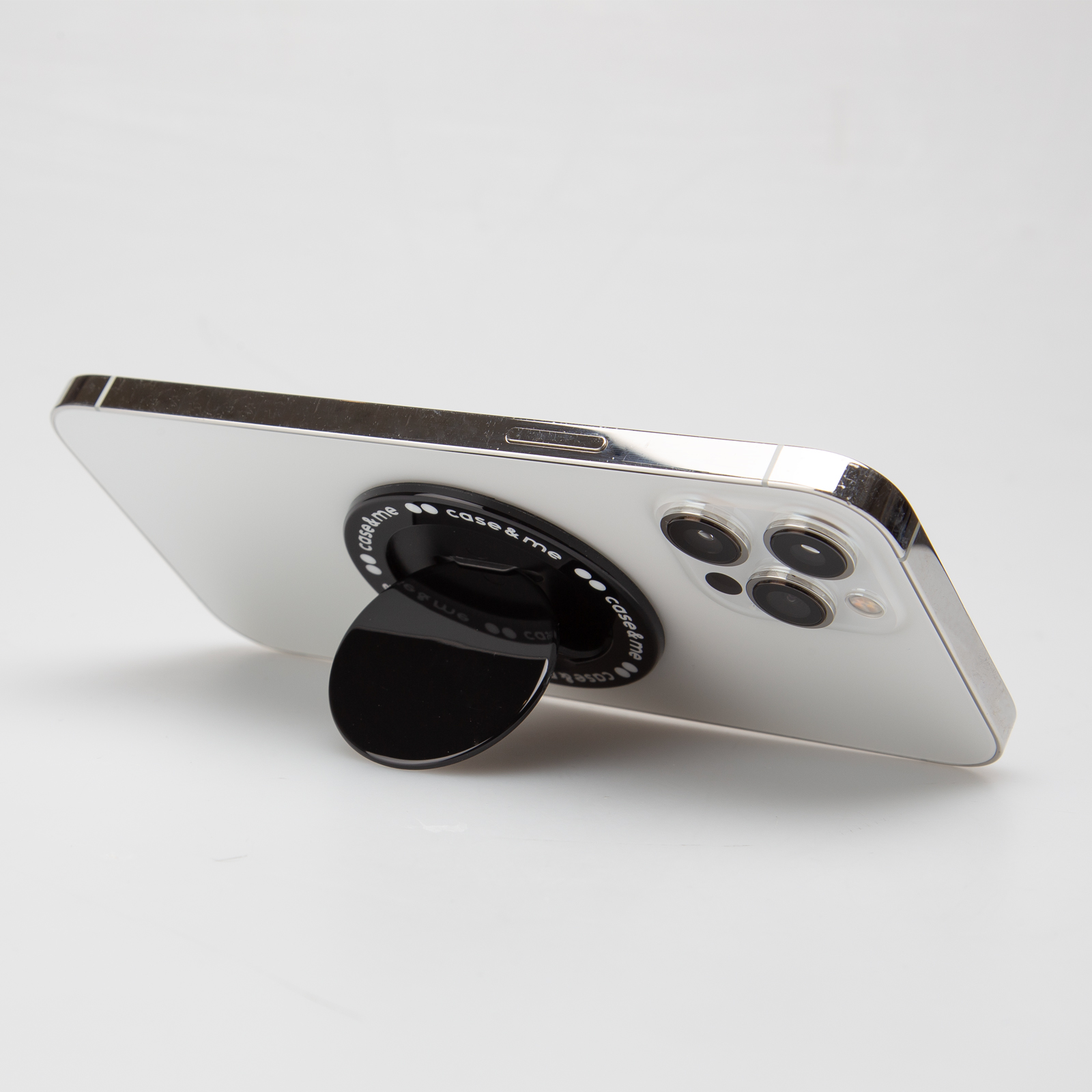 Un smartphone blanco con tres lentes de cámara descansa sobre una superficie blanca, sostenido por un soporte negro redondo.