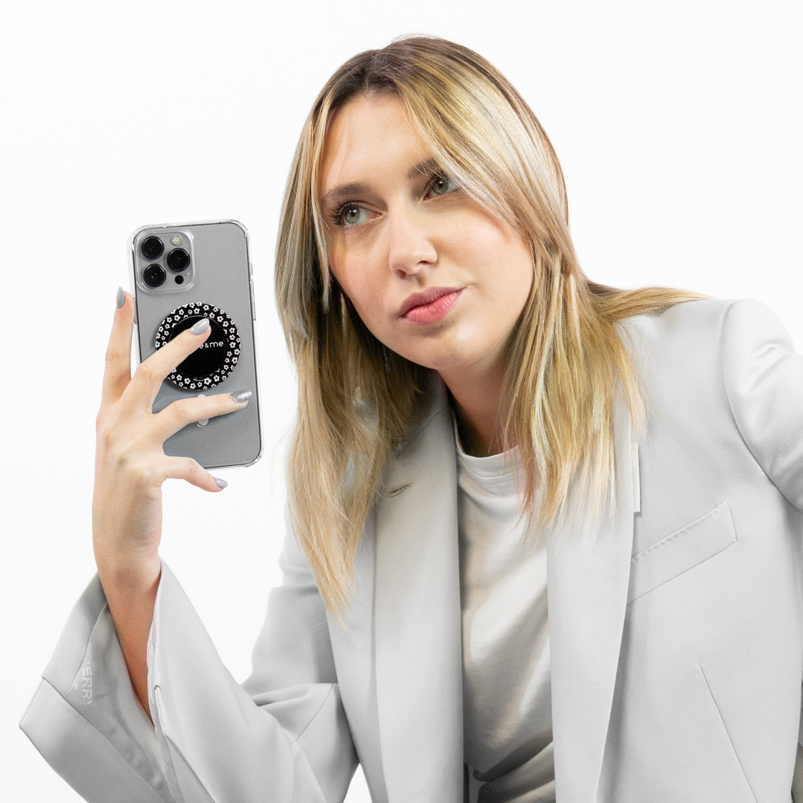 Una mujer rubia con un blazer gris sostiene con orgullo su smartphone con un llamativo soporte de anillo.