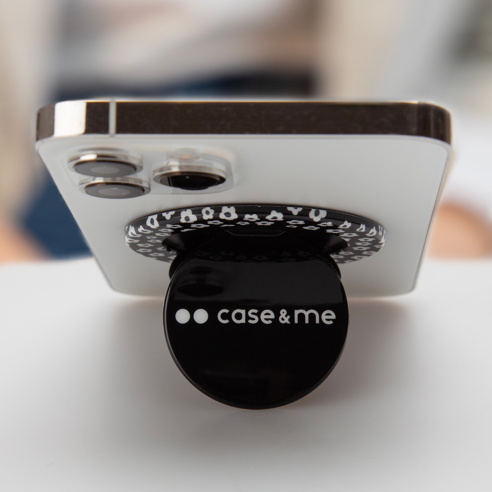 Un teléfono blanco en un soporte con logo negro 'case&me' sobre superficie blanca.