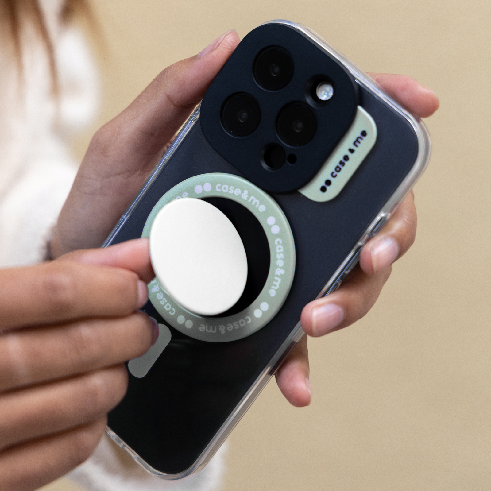 Persona sosteniendo un smartphone negro con un soporte verde pastel y un accesorio redondo blanco en la parte trasera.