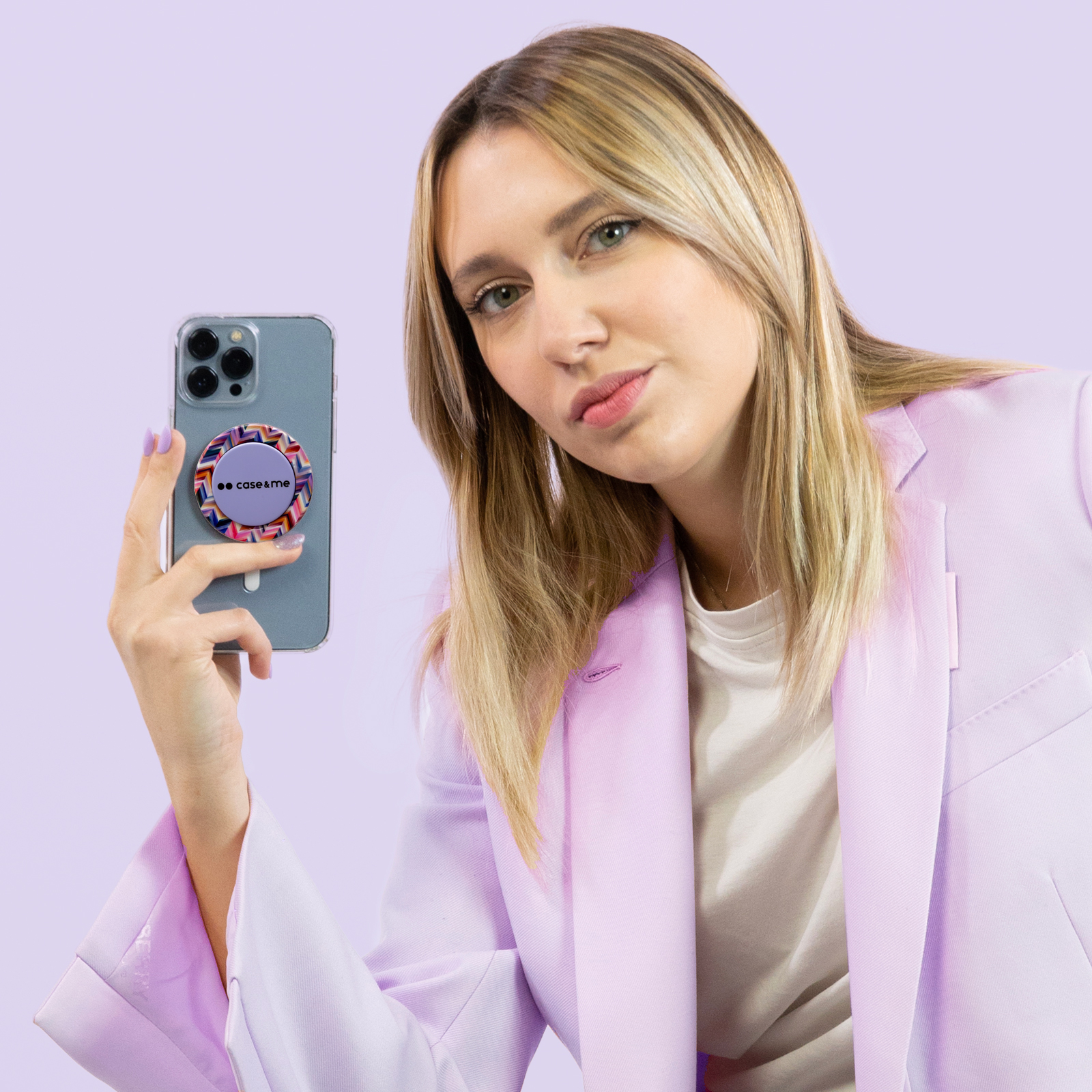 Mujer rubia con blazer morado sostiene un smartphone con soporte contra fondo morado.