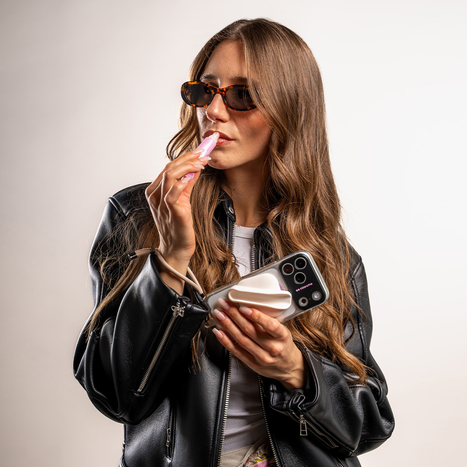 Una donna con giacca di pelle e occhiali da sole mette il lucidalabbra tenendo uno smartphone.