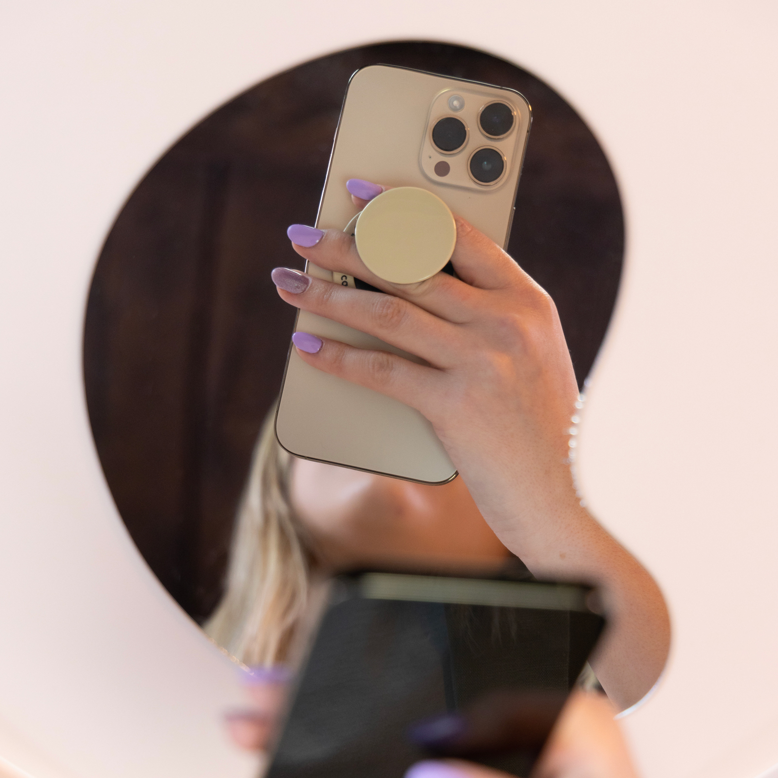 Persona con esmalte de uñas morado sostiene un smartphone dorado con popsocket en un espejo.