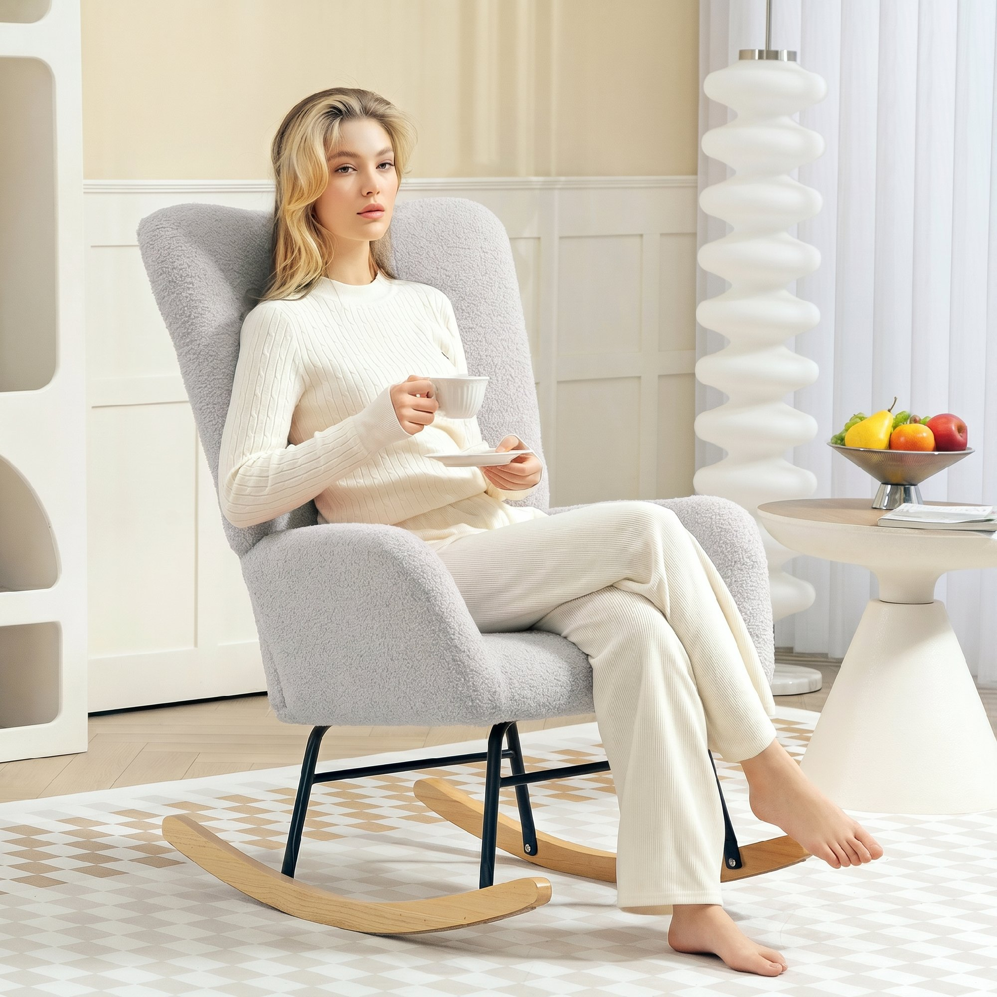 Una mujer rubia se sienta en una mecedora gris, sosteniendo una taza de té en una habitación luminosa.