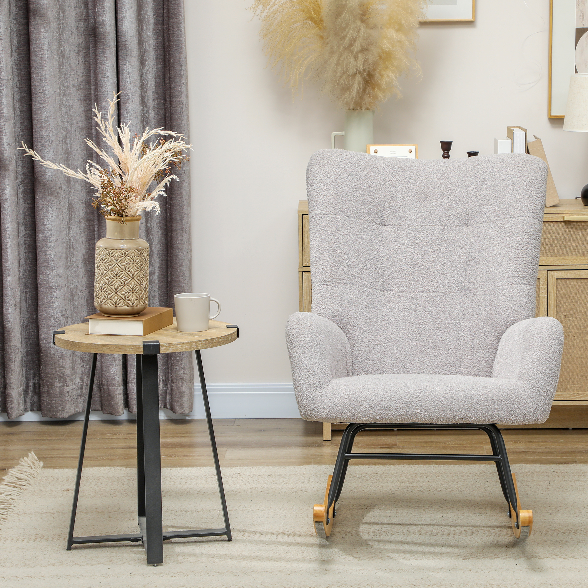 Una mecedora gris junto a una mesa redonda de madera con jarrón y taza, frente a cortinas grises.