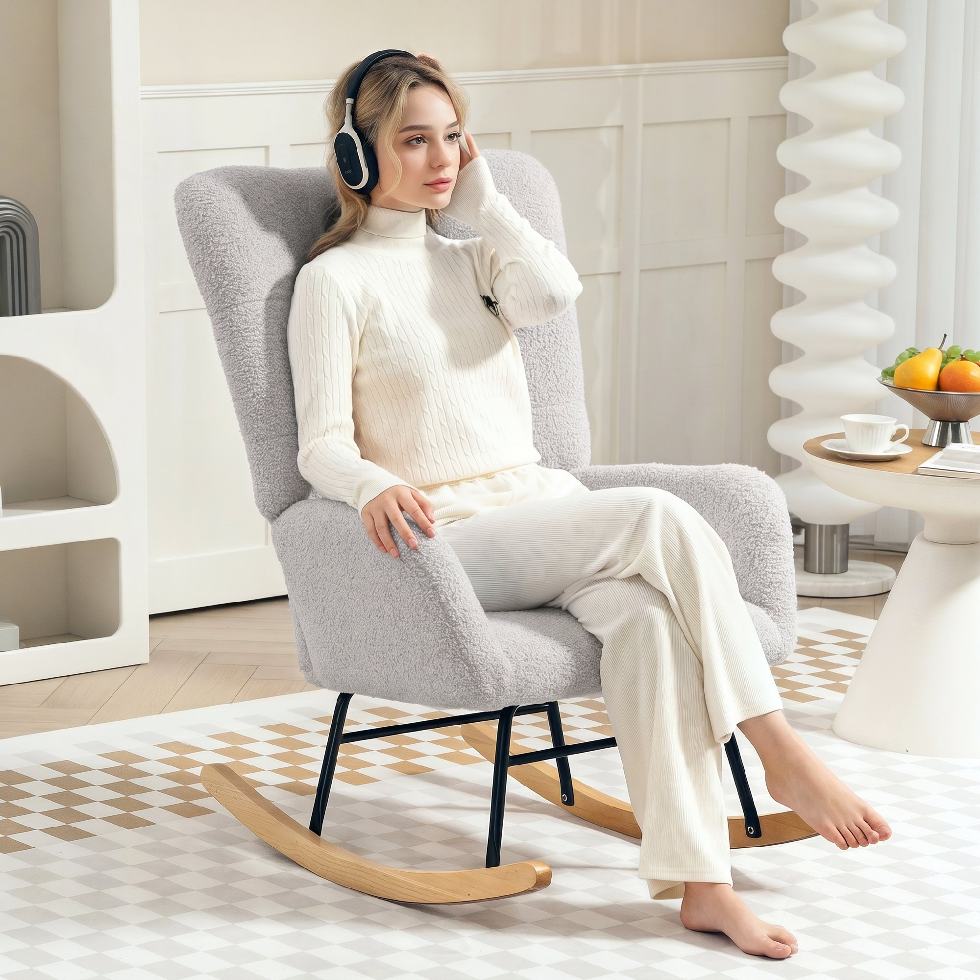 Una mujer rubia con ropa blanca se sienta en una mecedora gris, escuchando música con auriculares.