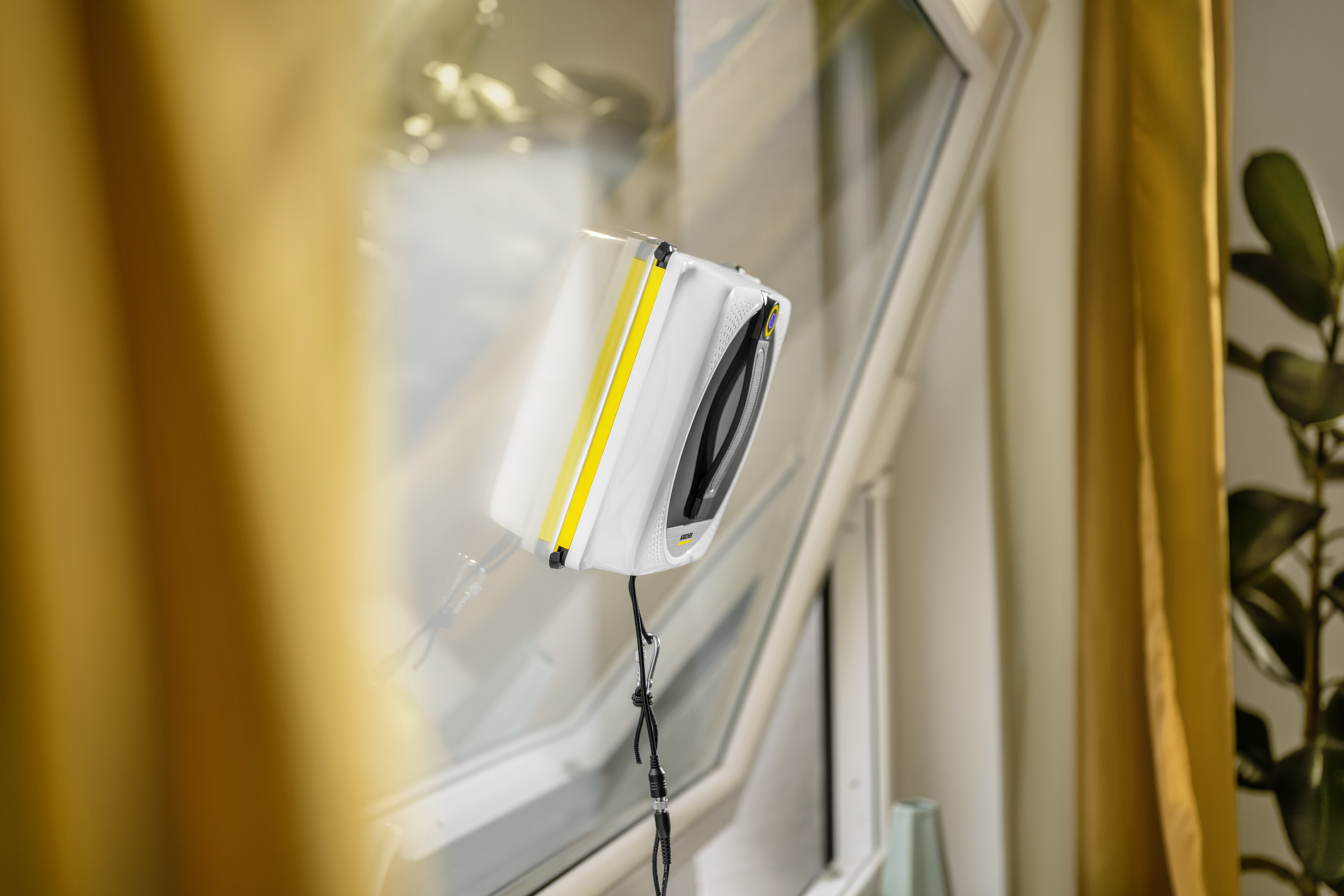 Un robot pulisci vetri bianco e giallo pulisce il vetro di una finestra inclinata in un interno con tende gialle.