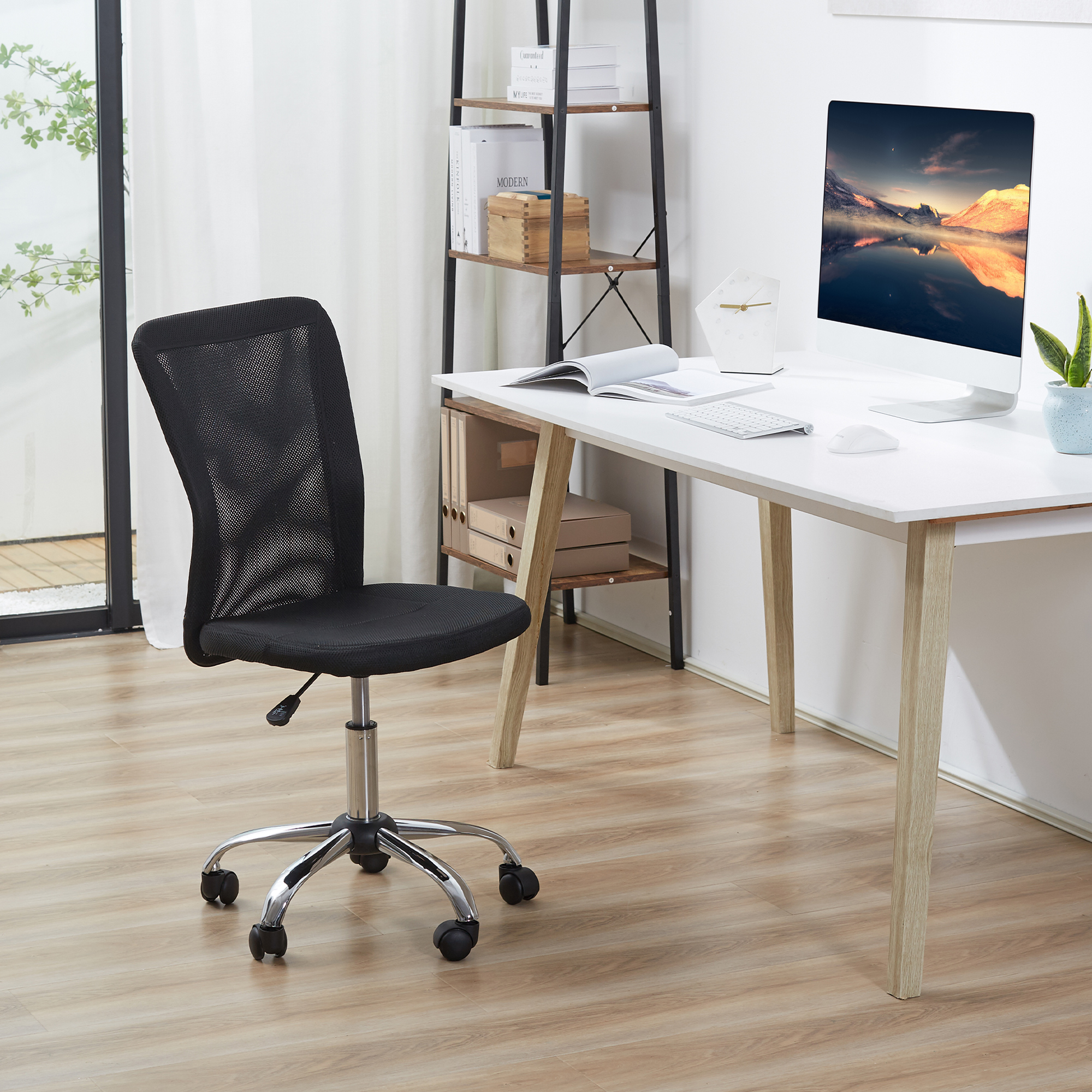 Una sedia da ufficio a rete nera si trova davanti a una scrivania bianca con monitor, tastiera e libro.