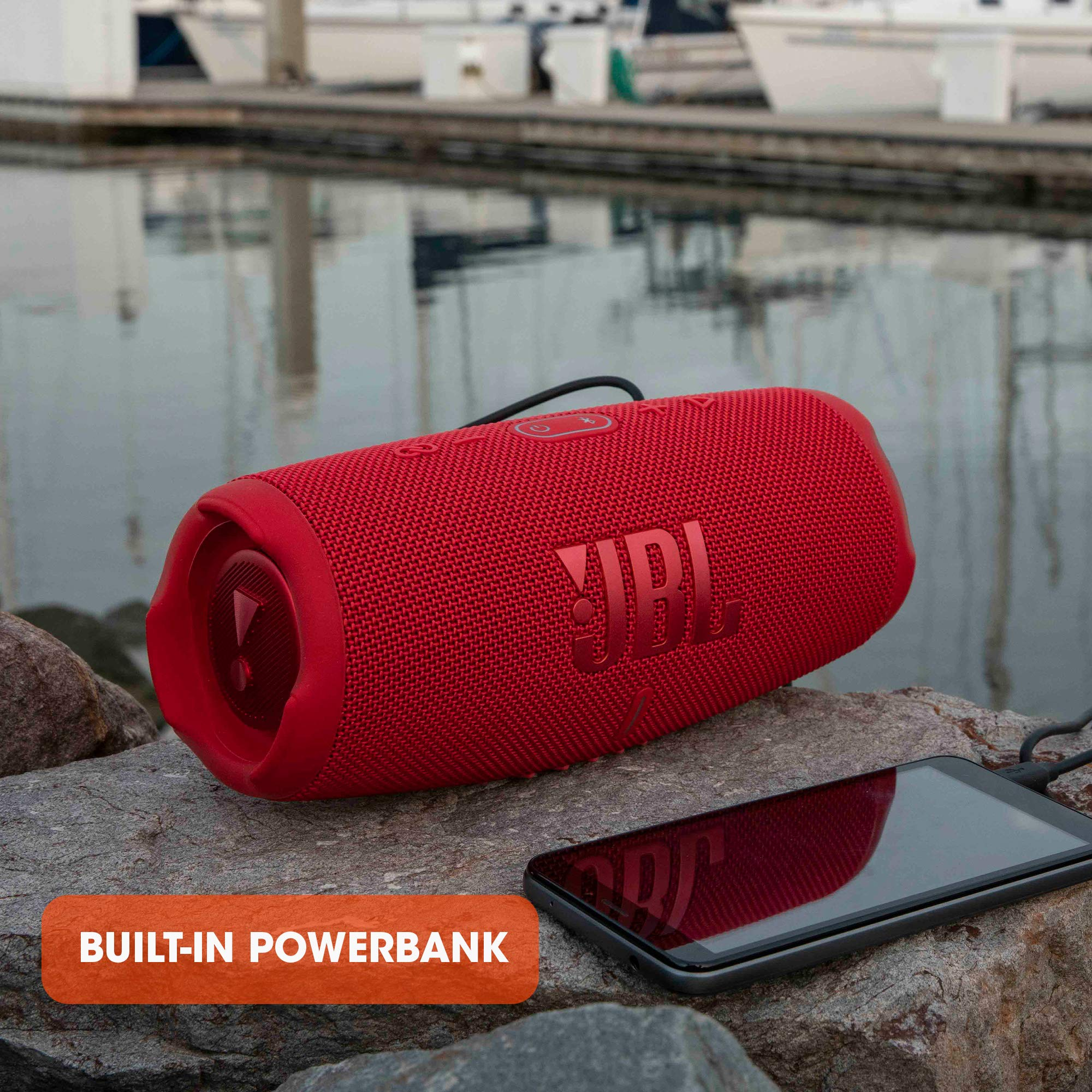 Haut-parleur JBL rouge et smartphone sur roche au bord de l'eau, texte : Built-in Powerbank.