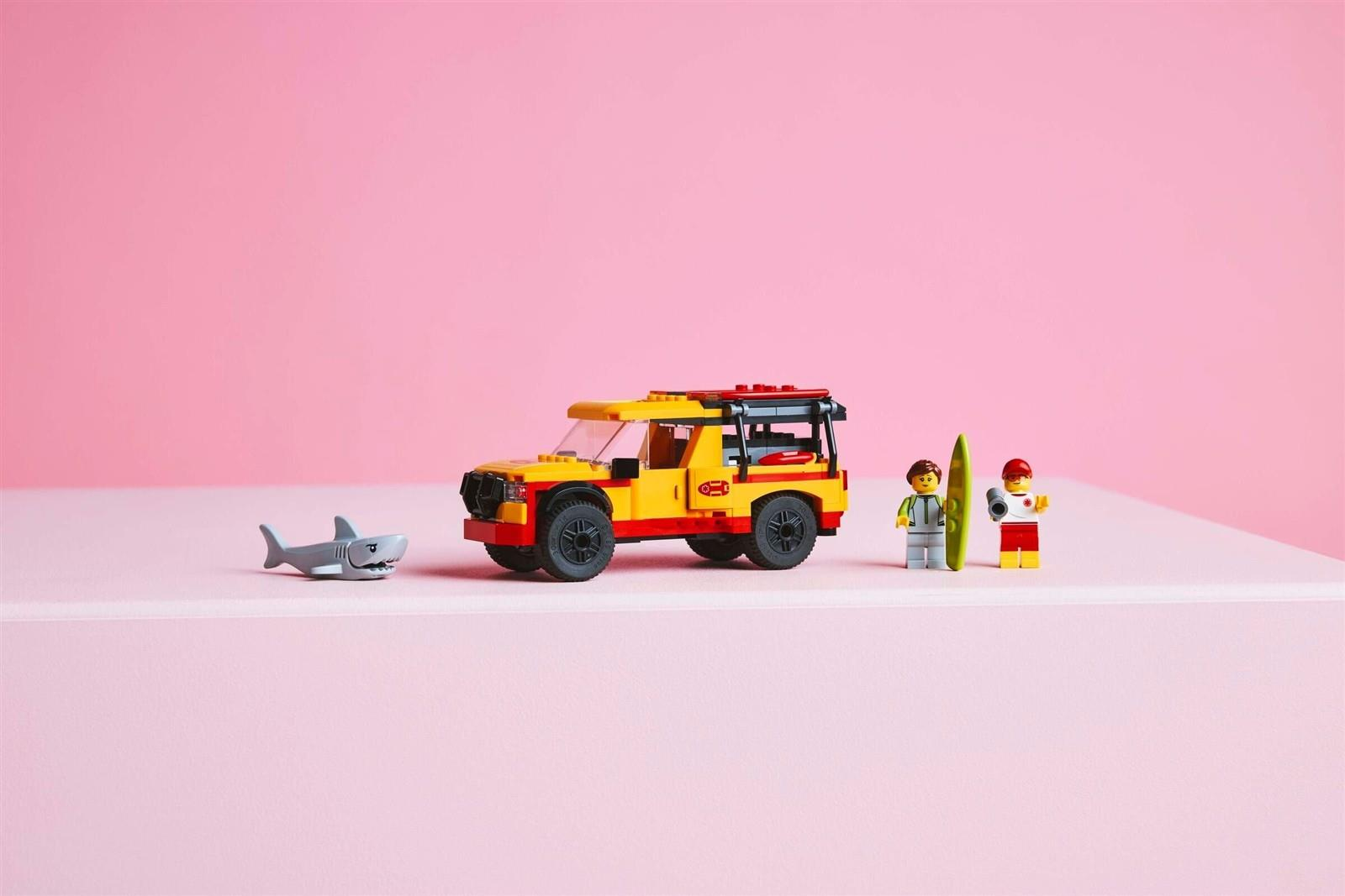 Lego-Strandrettungsset mit gelbem Lastwagen, zwei Figuren, Surfbrett und Haifisch auf rosa Hintergrund.
