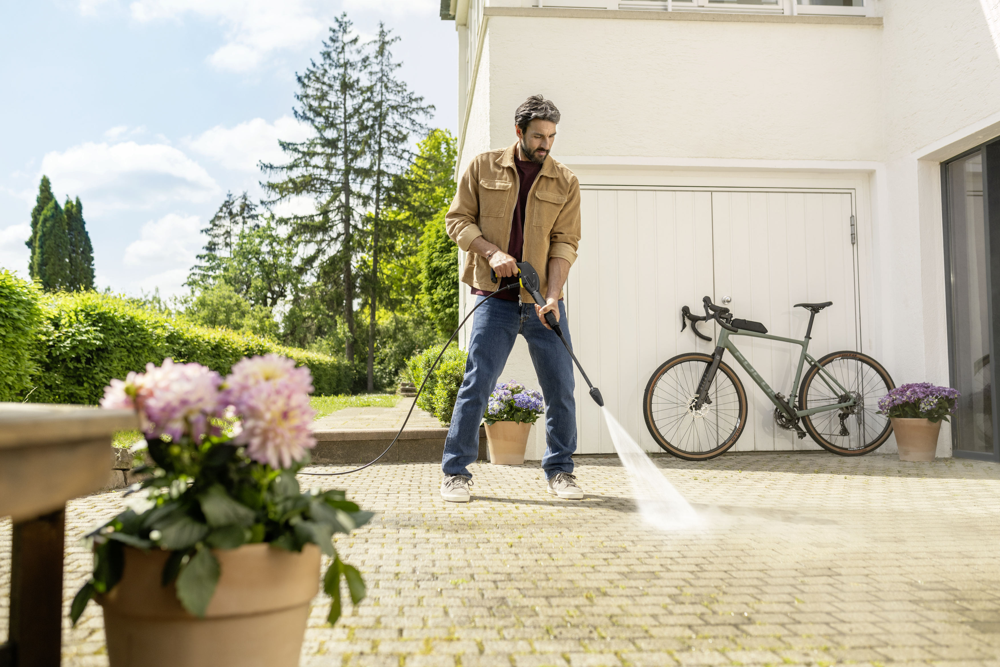 Ein Mann reinigt mit einem Hochdruckreiniger eine gepflasterte Terrasse vor einem Haus mit Fahrrad.