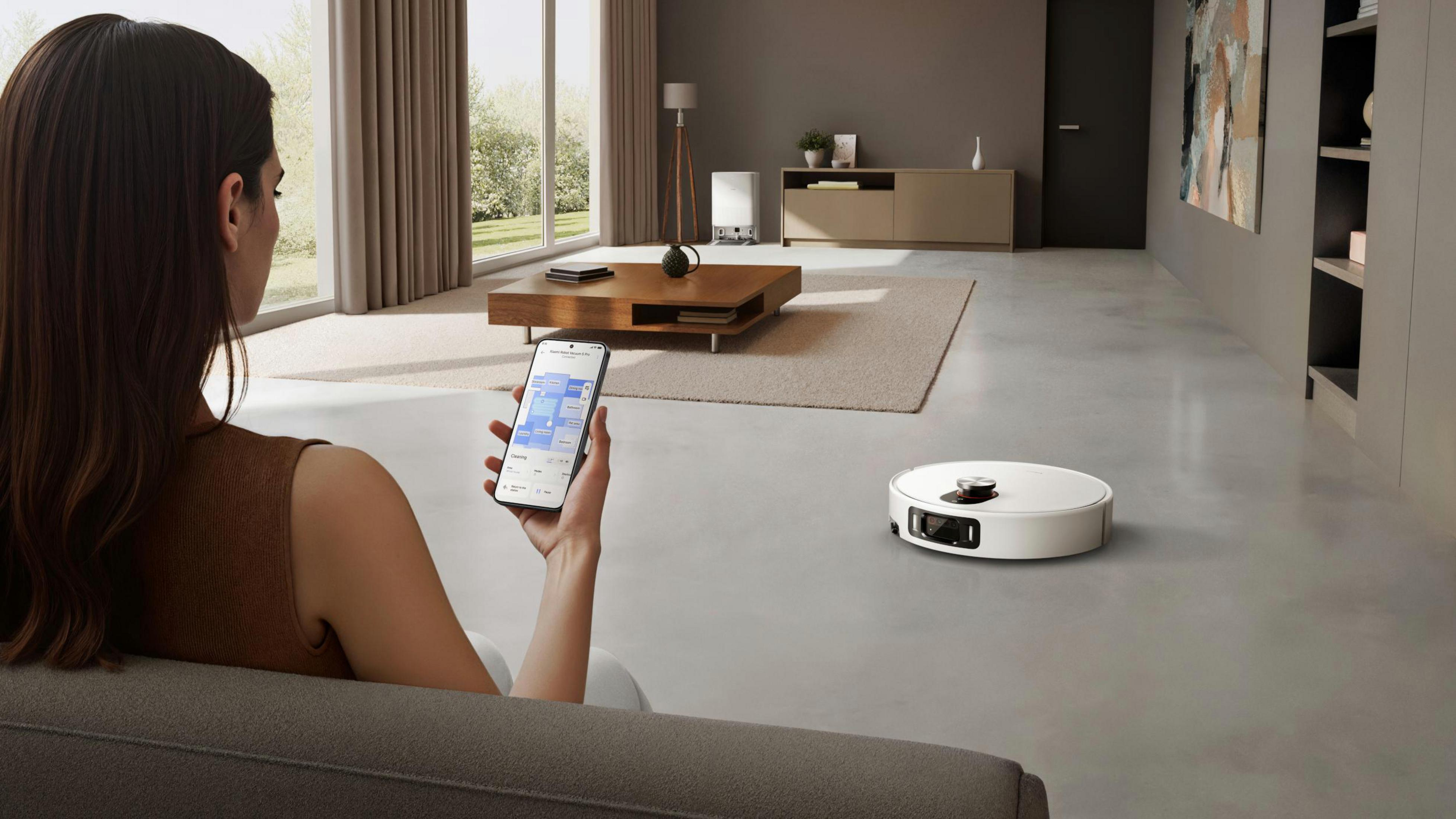 Kobieta siedzi na kanapie, sterując robotem sprzątającym za pomocą aplikacji w smartfonie w jasnym salonie.