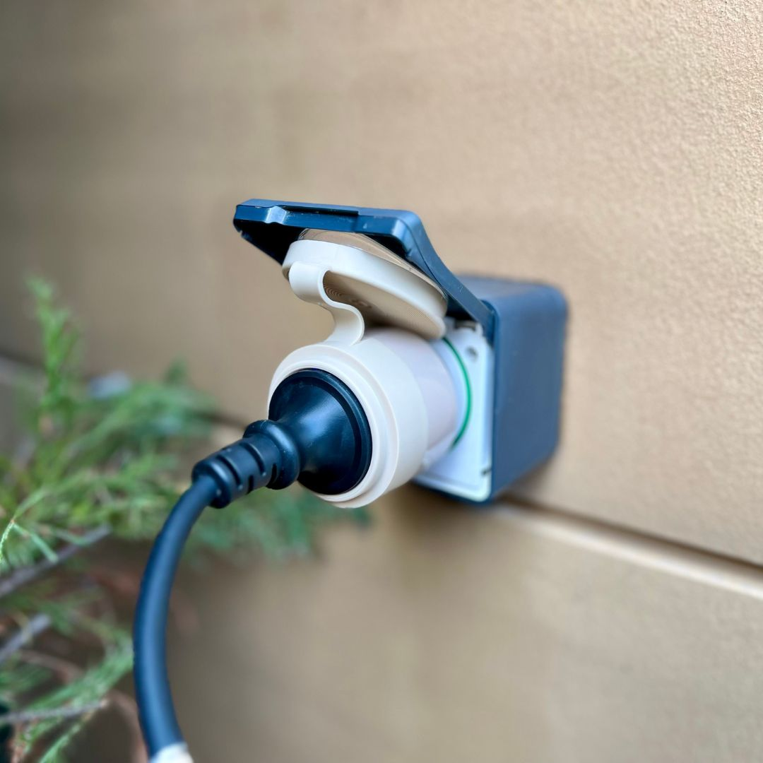 Eine wasserdichte Außensteckdose mit eingestecktem schwarzen Stecker ist an einer beigen Wand montiert.