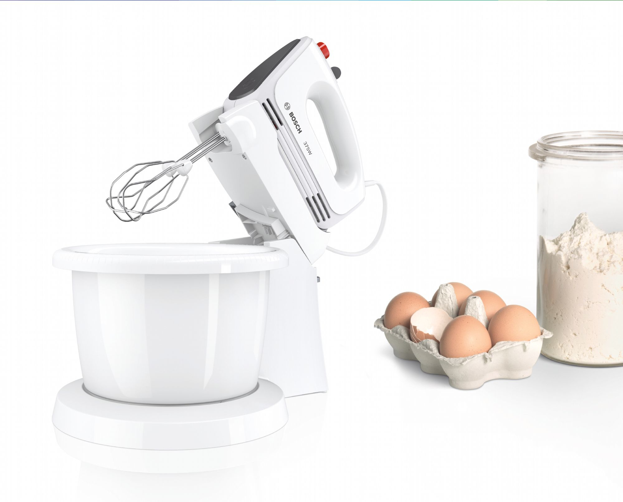 Un mixer a mano bianco Bosch con ciotola accanto a uova in cartone e un barattolo di farina.