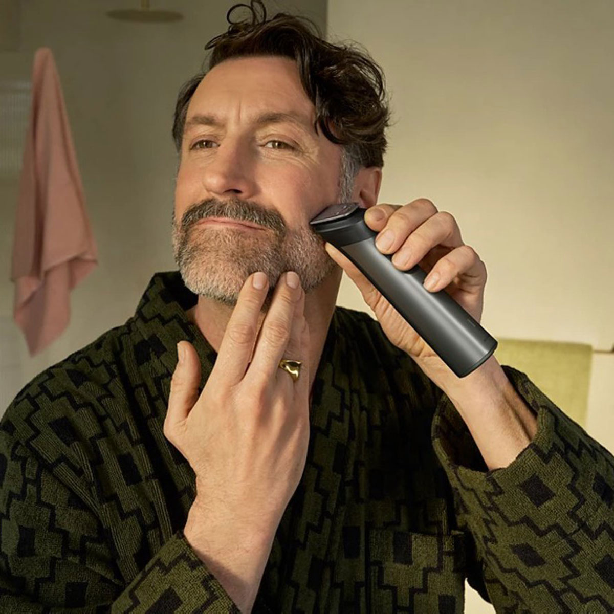 Un uomo con una vestaglia verde fantasia si rade la barba con un rasoio in bagno.