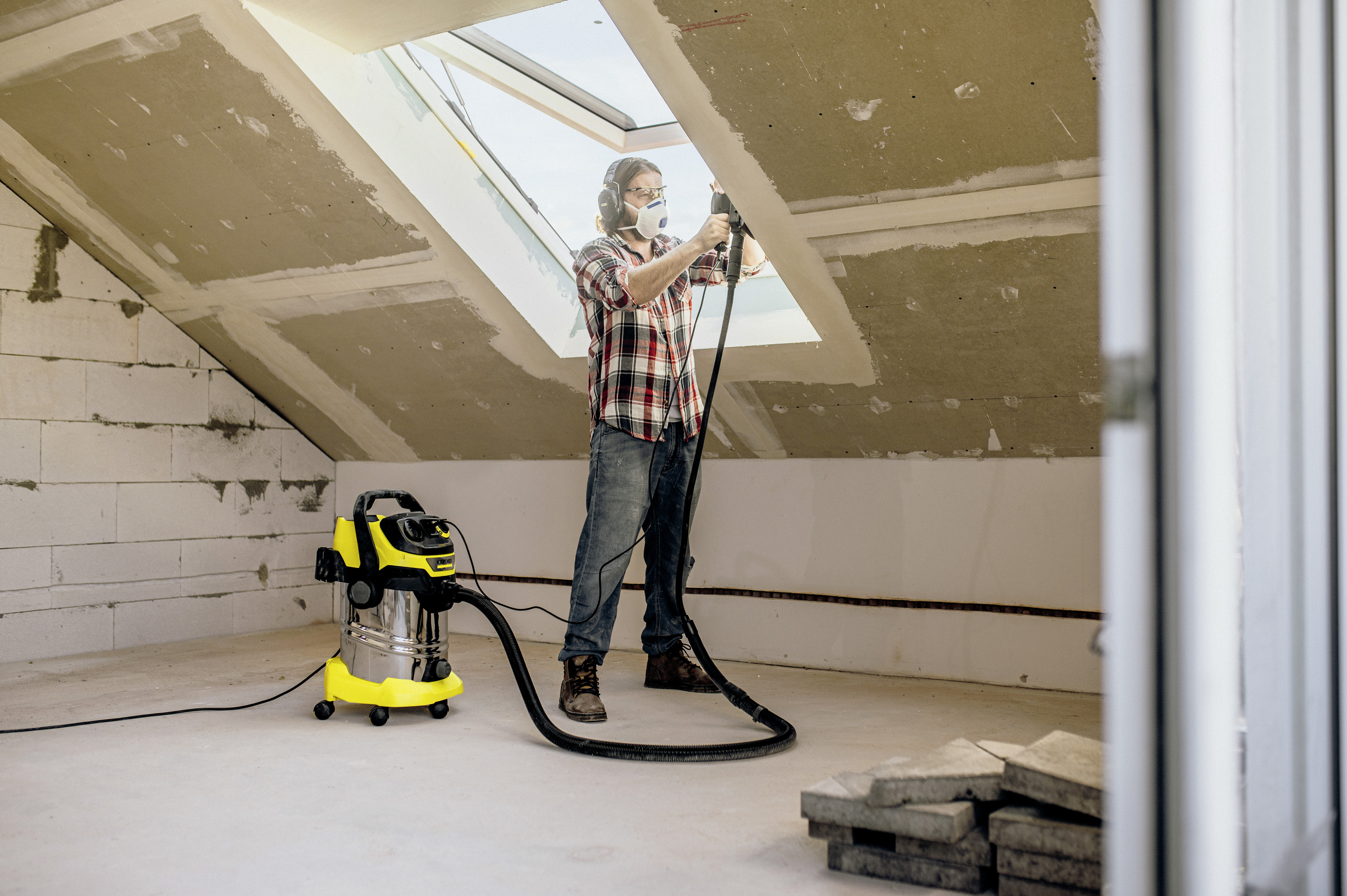 Handwerker mit Schutzkleidung nutzt ein Elektrowerkzeug und einen gelben Sauger in einem unfertigen Dachgeschoss.