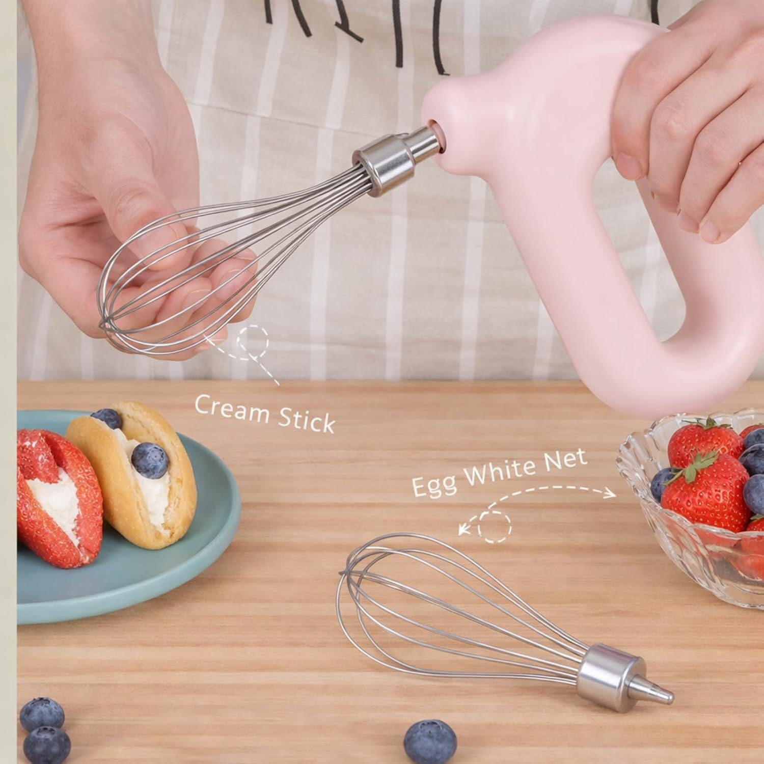 Eine Person hält einen rosa elektrischen Mixer mit Schneebesen, daneben Gebäck und eine Schüssel Obst.